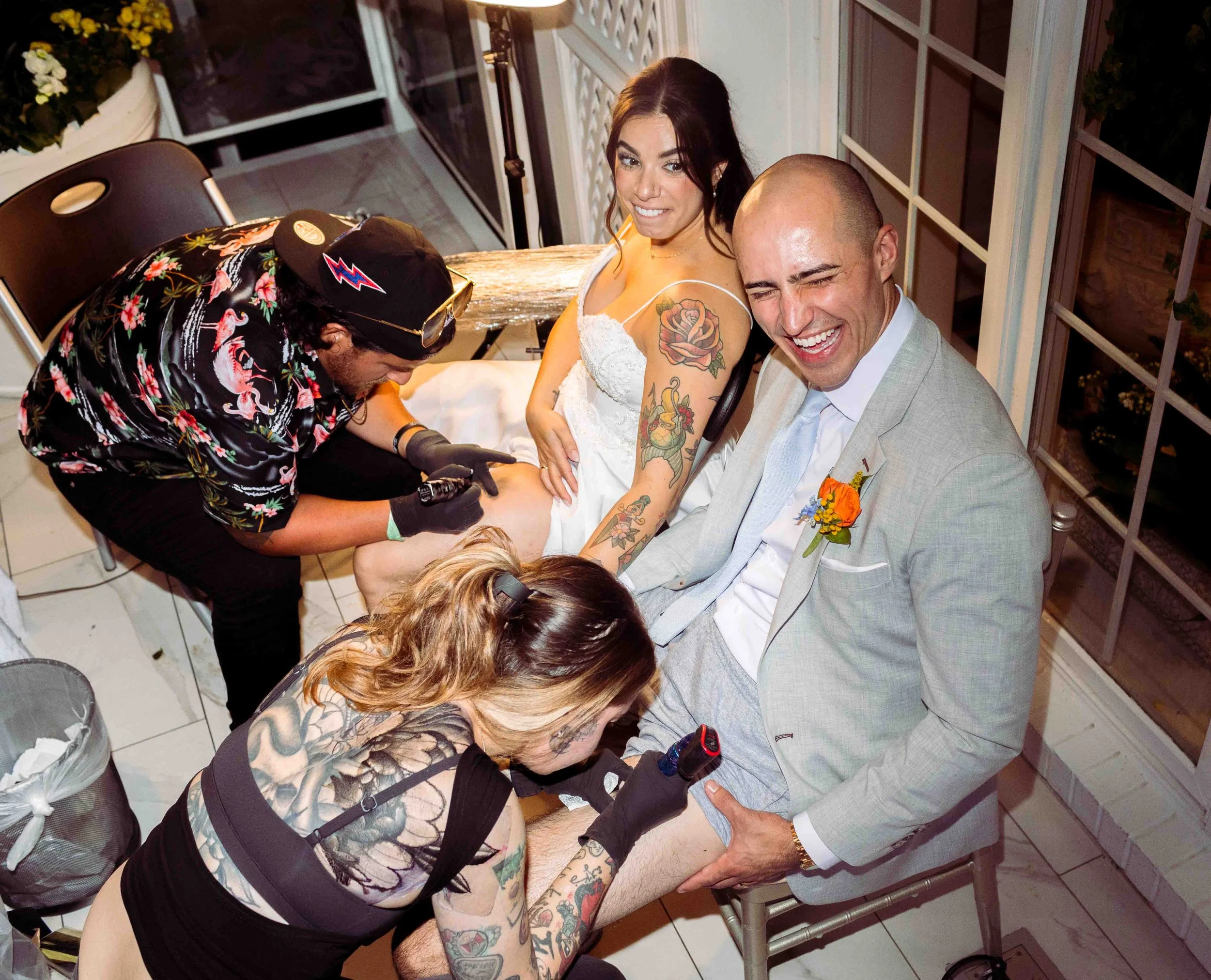 Tattoo artist tattooing a man's thigh at a wedding reception, with a smiling bride and groom watching.