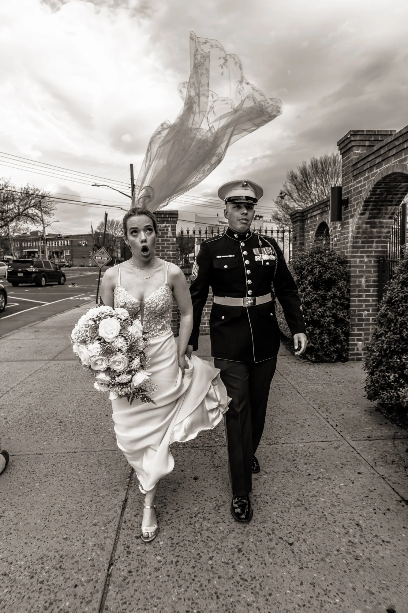 A bride in a wedding dress holding a bouquet of flowers, walking with a uniformed soldier outside near a brick gate. The bride has an expression of surprise or excitement, and a veil is flying in the air.