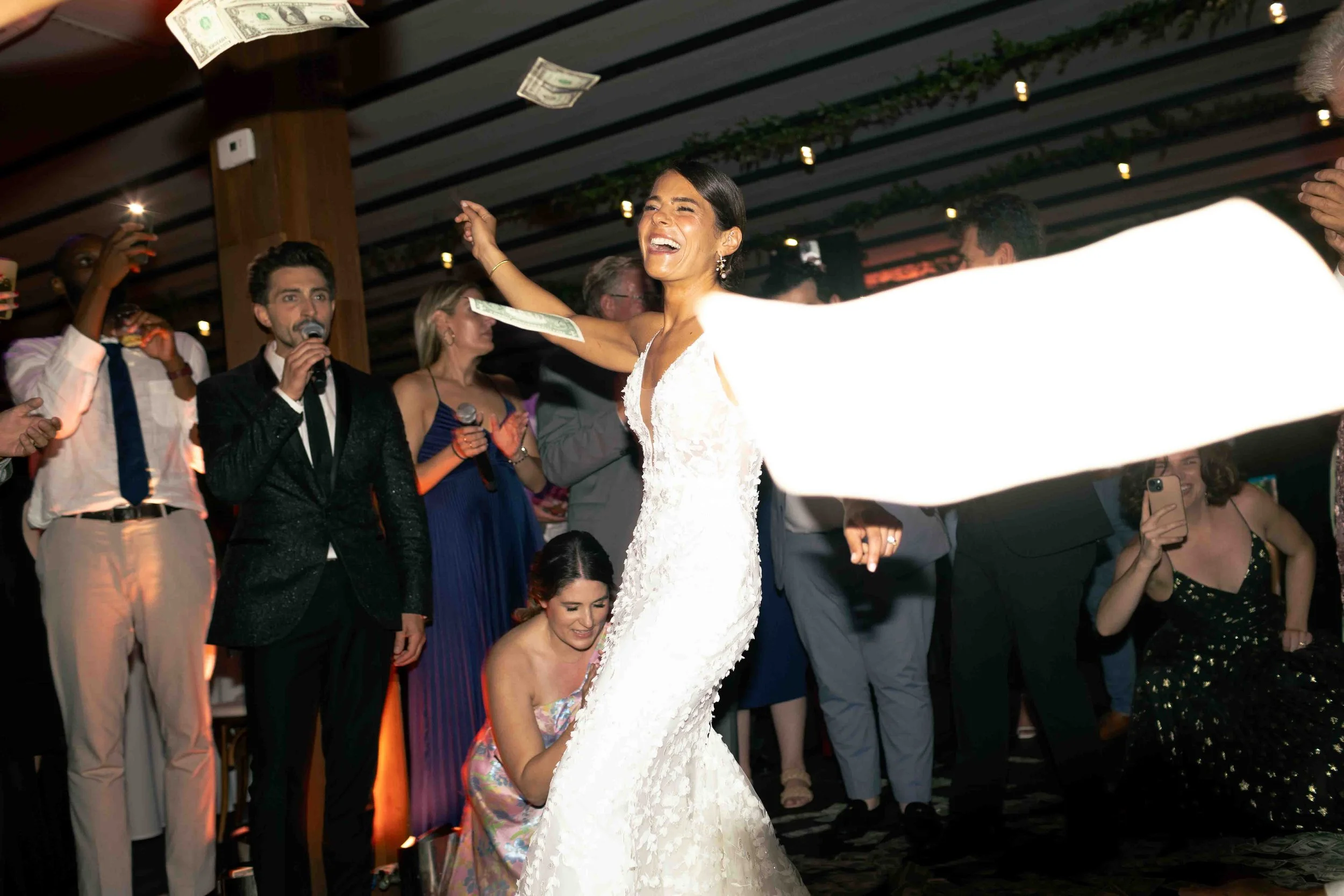 A woman in a white wedding dress dances joyfully surrounded by people at a wedding reception, with dollar bills being thrown in the air.