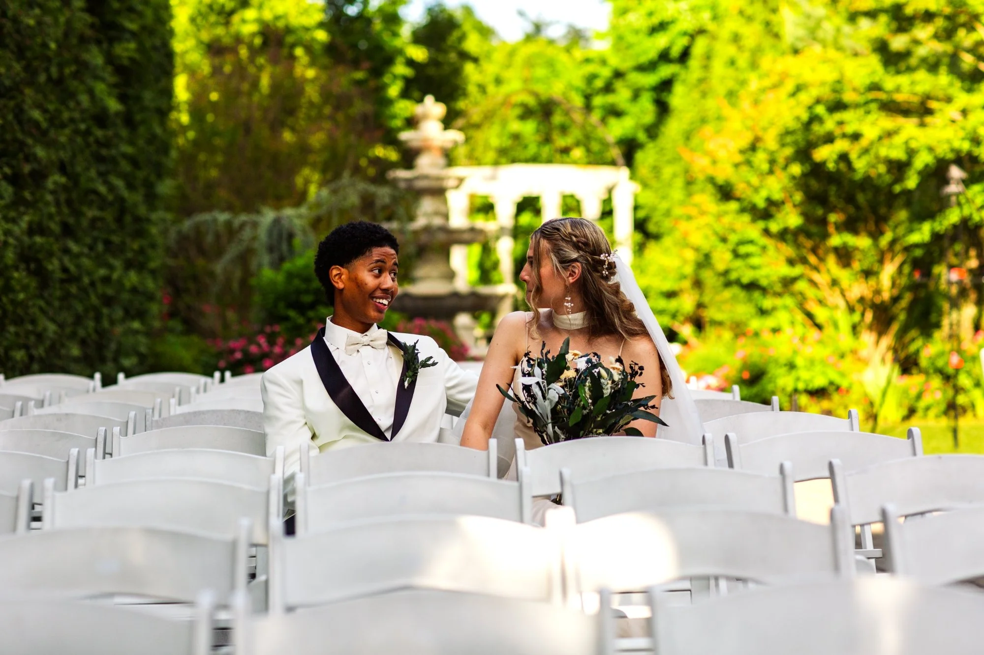 A bride and groom sitting together during a wedding ceremony outdoors on a sunny day, with greenery and flowers in the background.