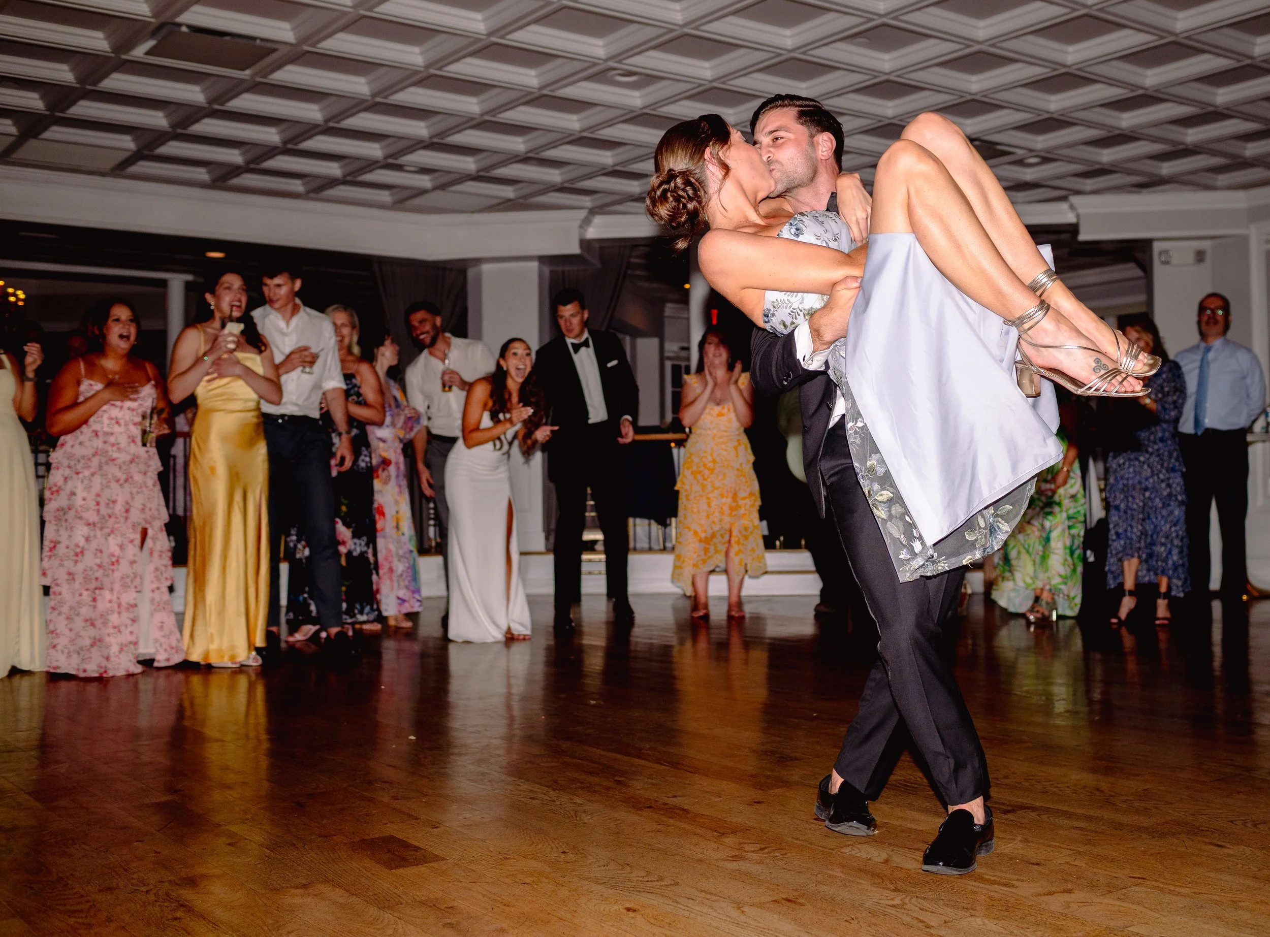Groom dancing and dipping bride during wedding reception as guests watch and cheer in elegant attire.