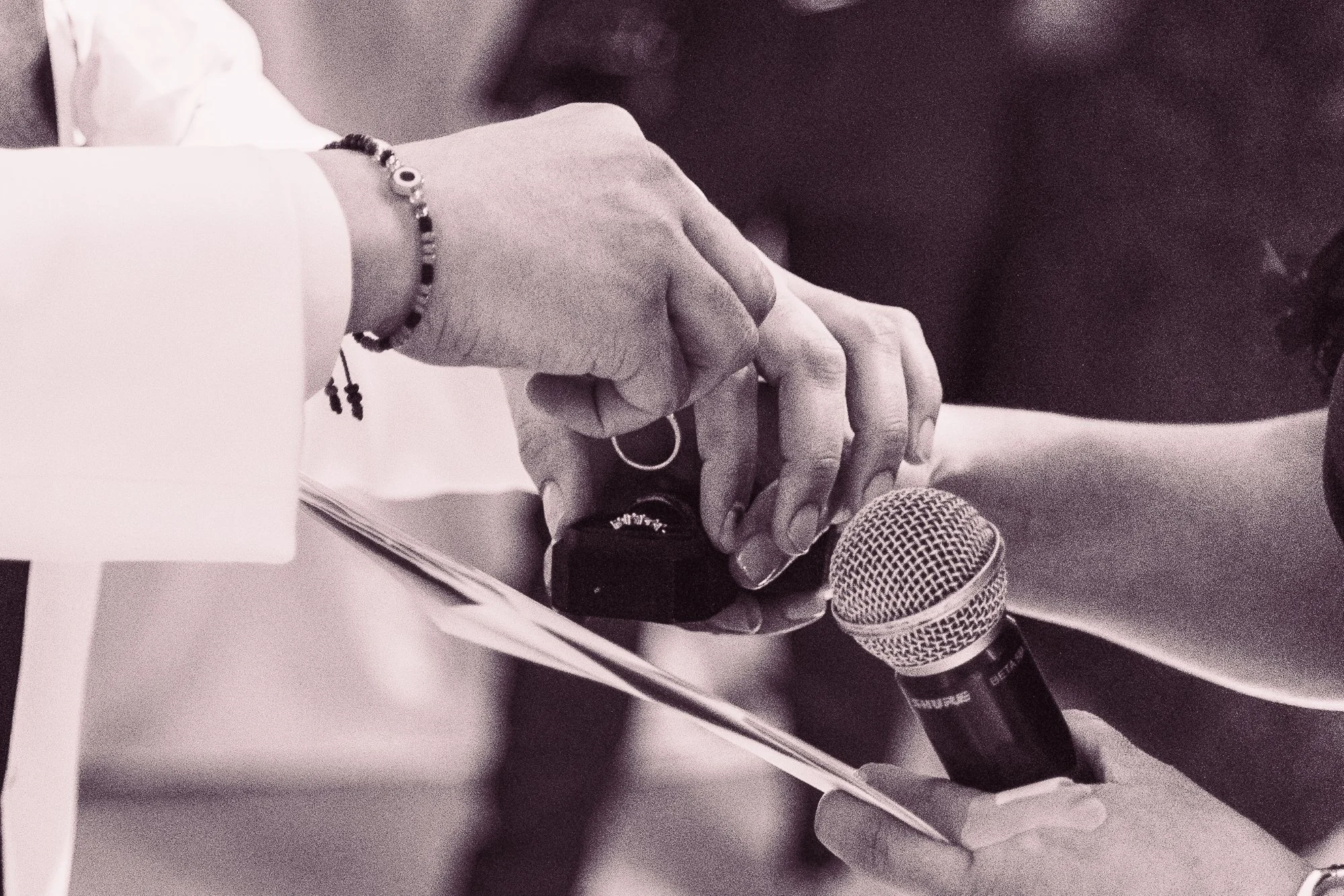 Close-up of a person holding a microphone in front of a piece of paper, with their hand displaying jewelry and rings, suggesting they are preparing to speak or give a presentation.