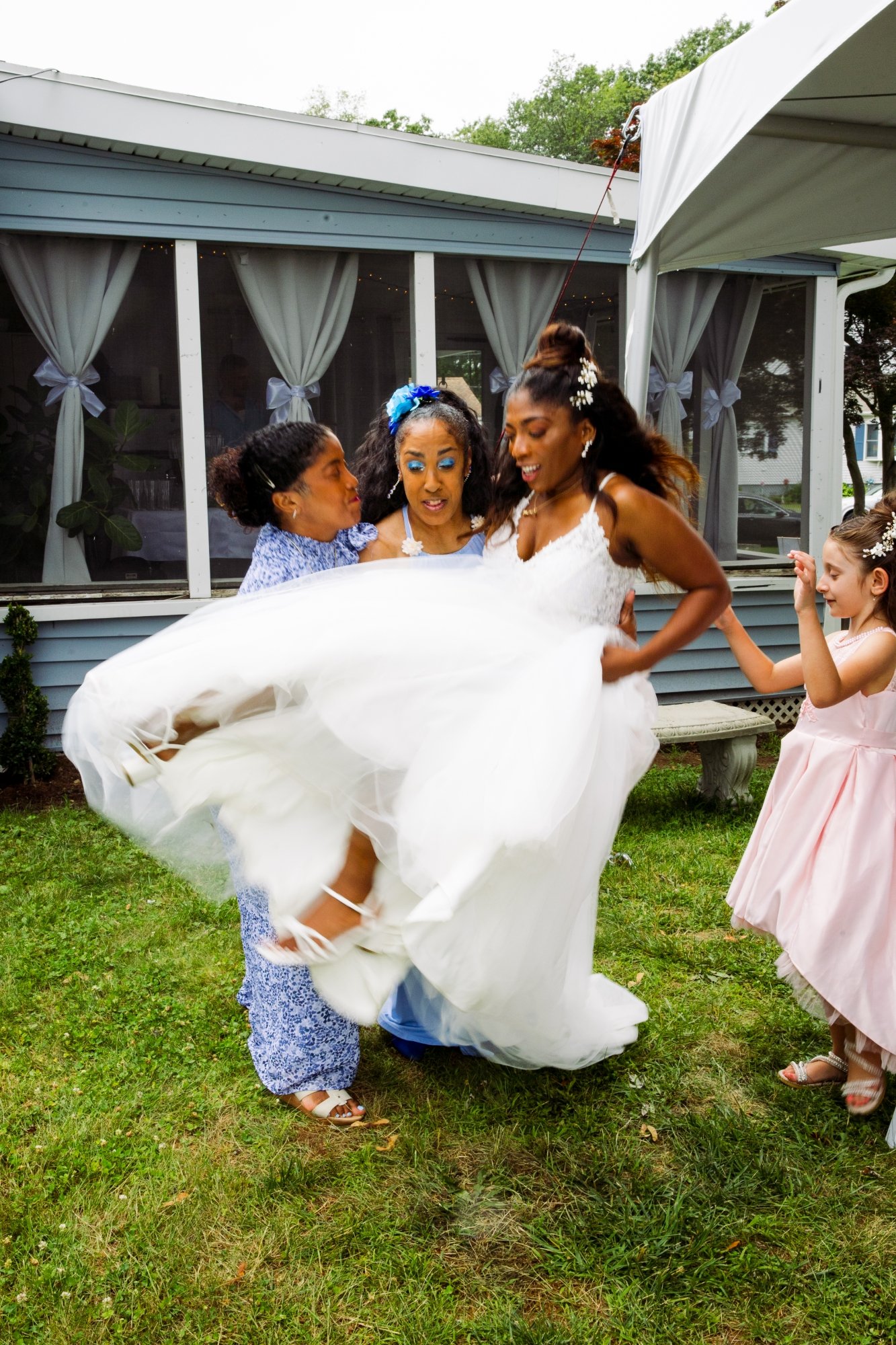 A bride wearing a white wedding dress and blue eye makeup surrounded by several women, one in a gray patterned dress and two young girls in pink dresses, enjoying a celebration outdoors in front of a house with light blue siding and large windows.