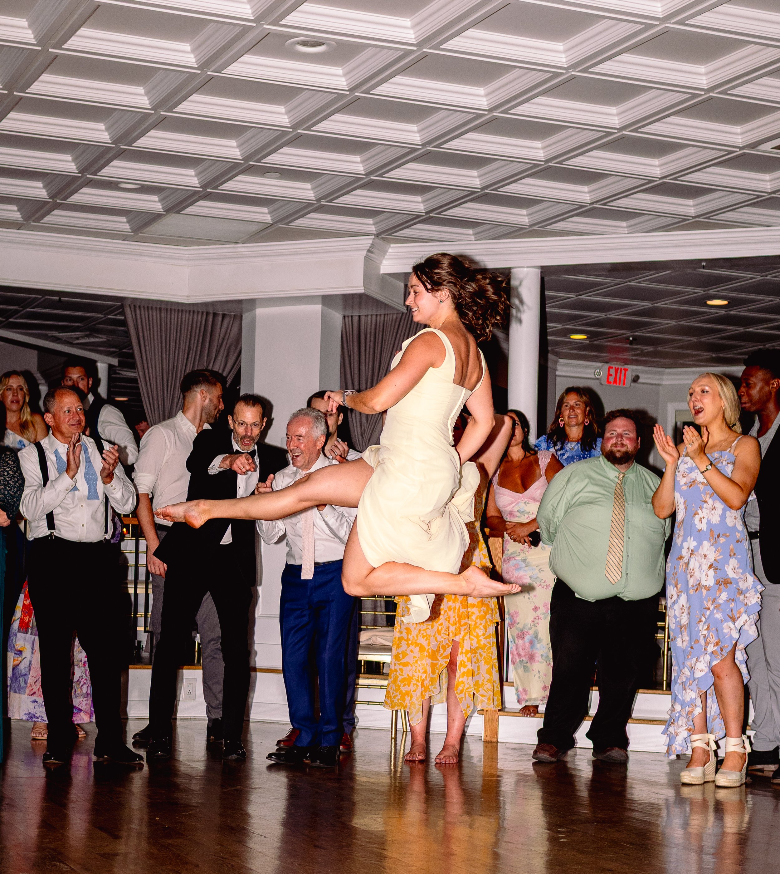 A woman in a pale yellow dress is being lifted and dancing on the floor of a lively party or celebration with many smiling and clapping guests around her.