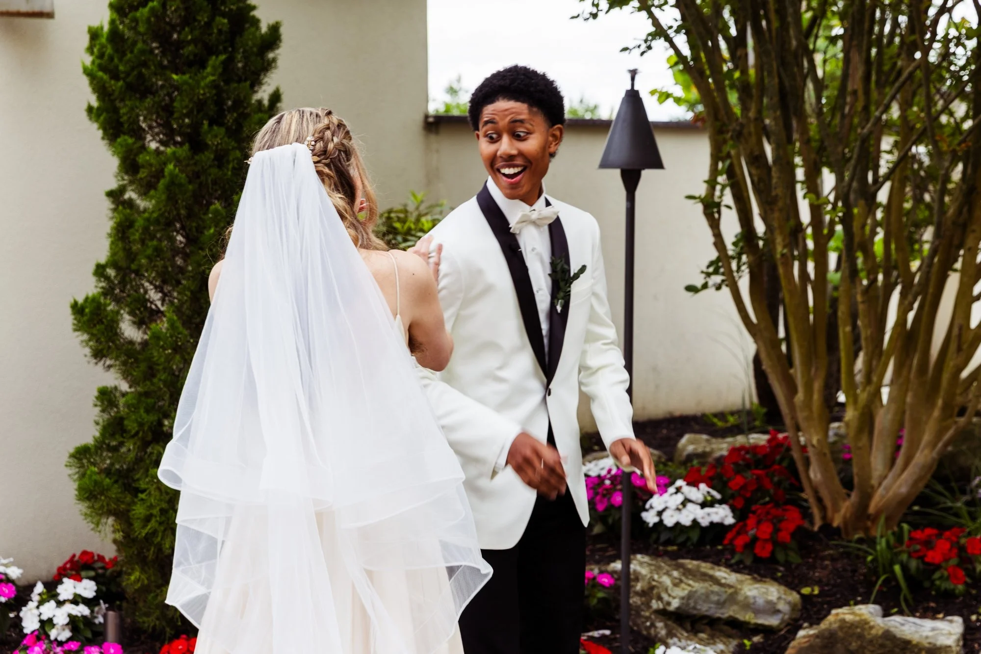 A happy bride and groom sharing a moment outdoors, with the bride wearing a white wedding dress and veil, and the groom in a white tuxedo jacket with a black bow tie, surrounded by colorful flowers and greenery.