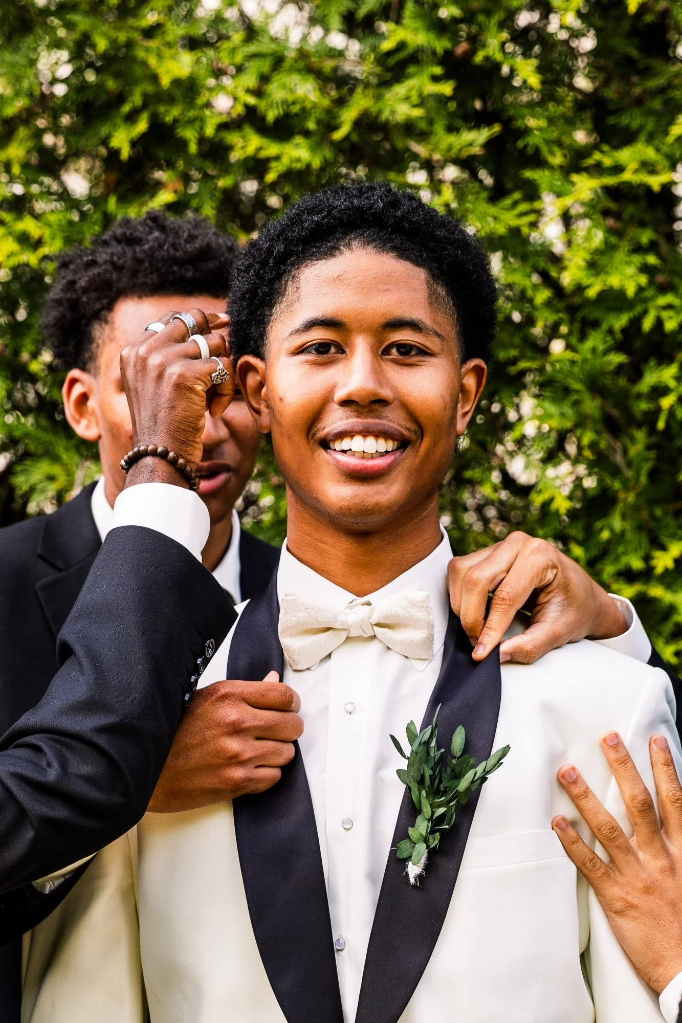 A young man in a white tuxedo with a black lapel and a white bow tie smiling at the camera, standing outdoors surrounded by greenery, with hands adjusting his tuxedo. Another man, partially visible, stands behind him, touching his shoulder.