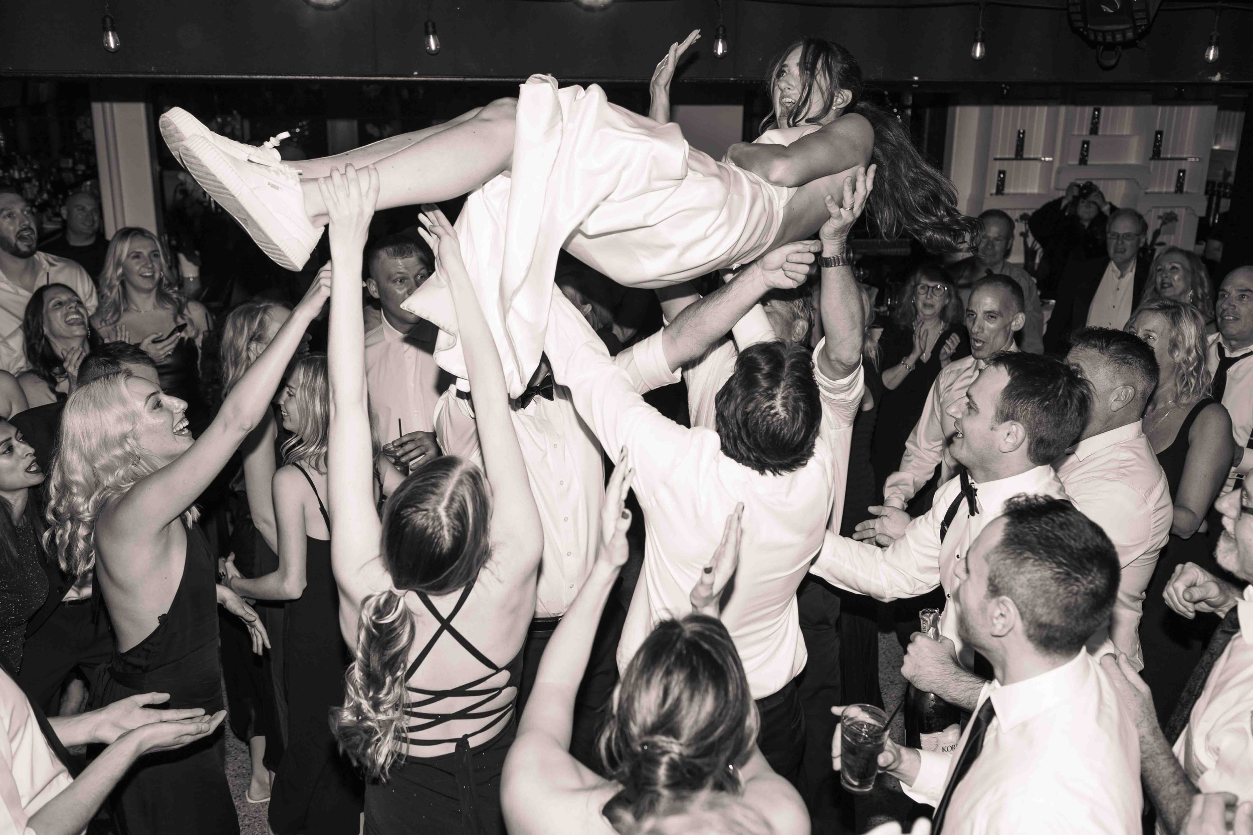 People at a celebration lift a woman in a white dress above their heads, smiling, with a lively crowd in the background.