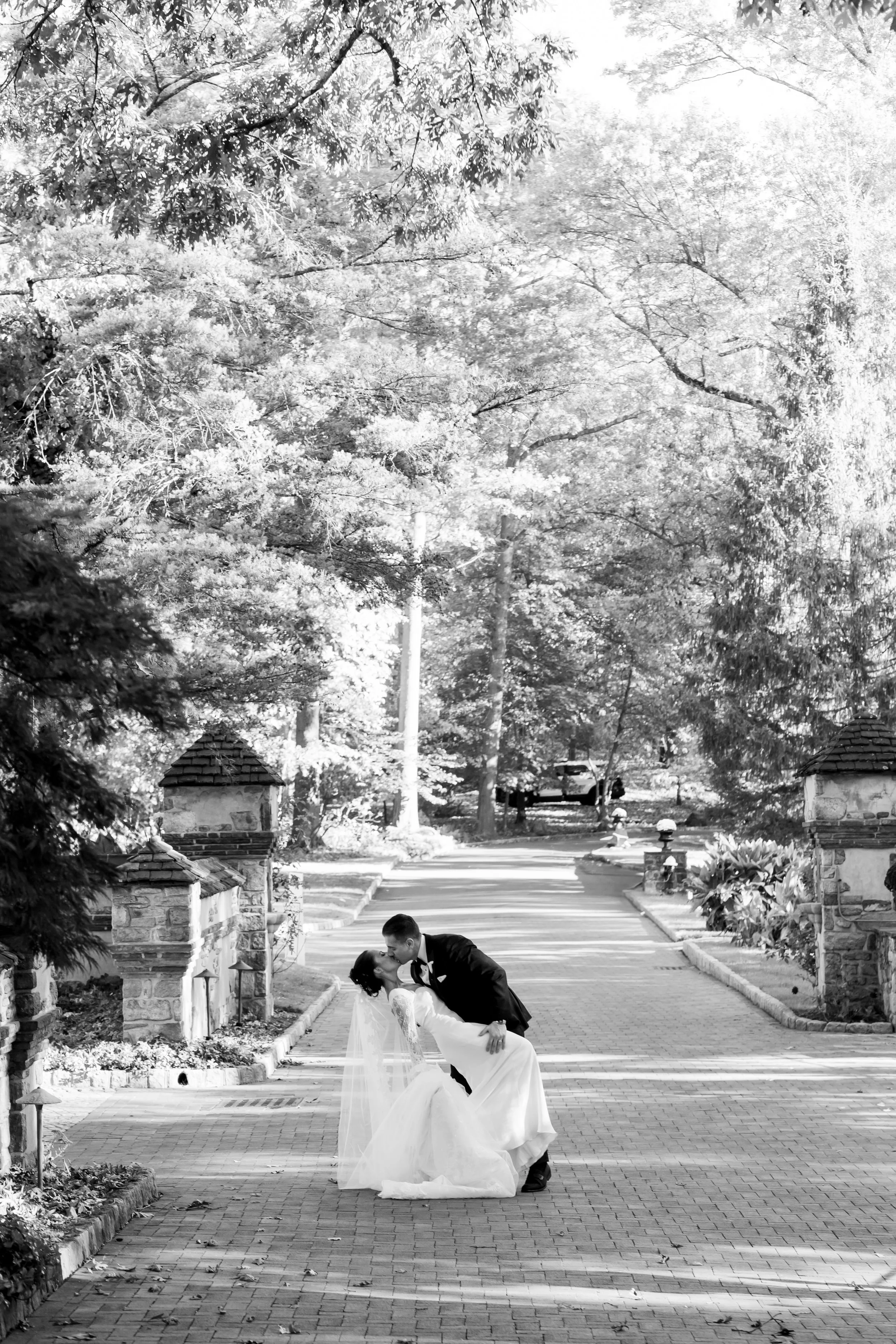 A black and white photo of a newlywed couple dancing outdoors on a brick pathway surrounded by trees and stone pillars.