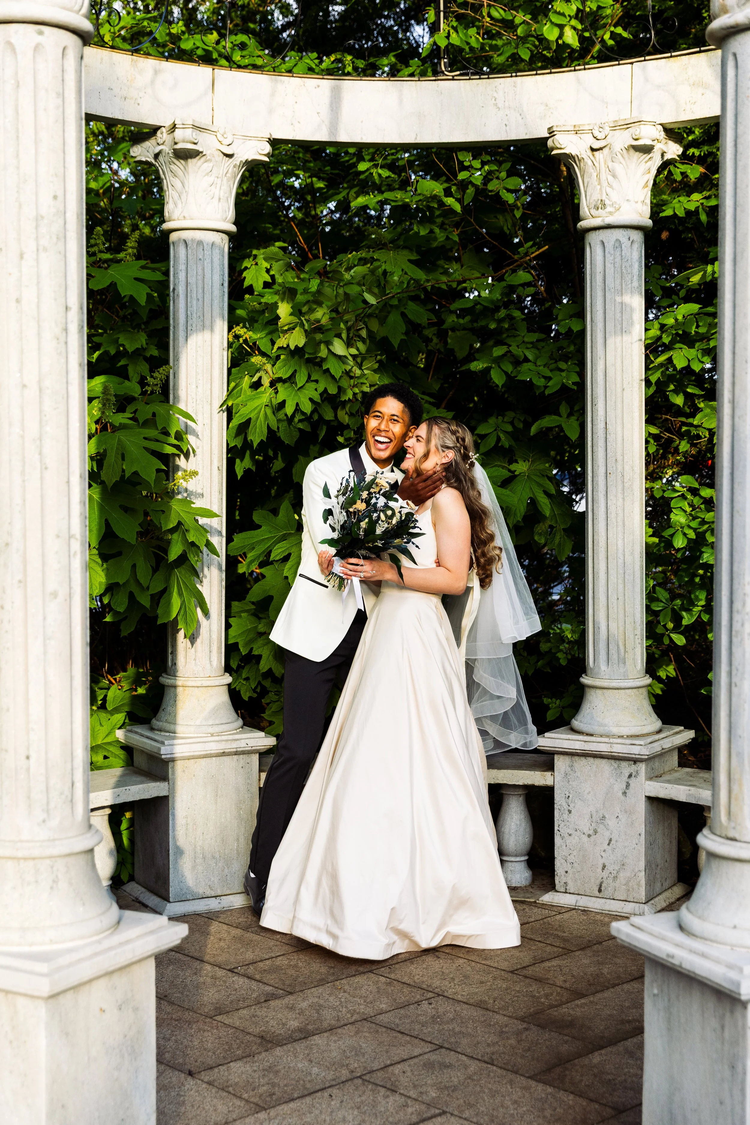 A happy interracial couple in wedding attire standing under a classical marble gazebo with green foliage in the background, sharing an intimate moment and laughing.