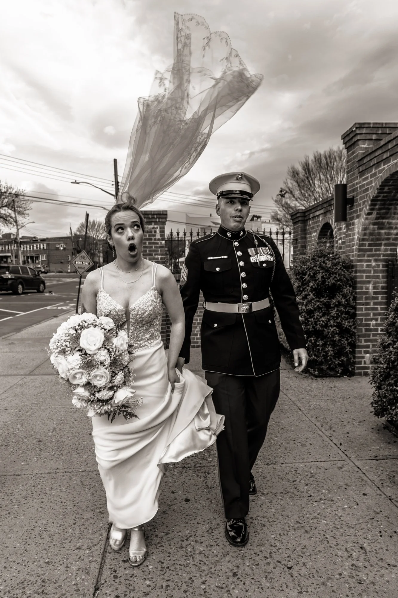 A wedding scene in black and white showing a bride in a gown holding a bouquet of flowers walking outdoors next to a man in military uniform. The bride looks surprised with her mouth open, and her veil is flying above her head.