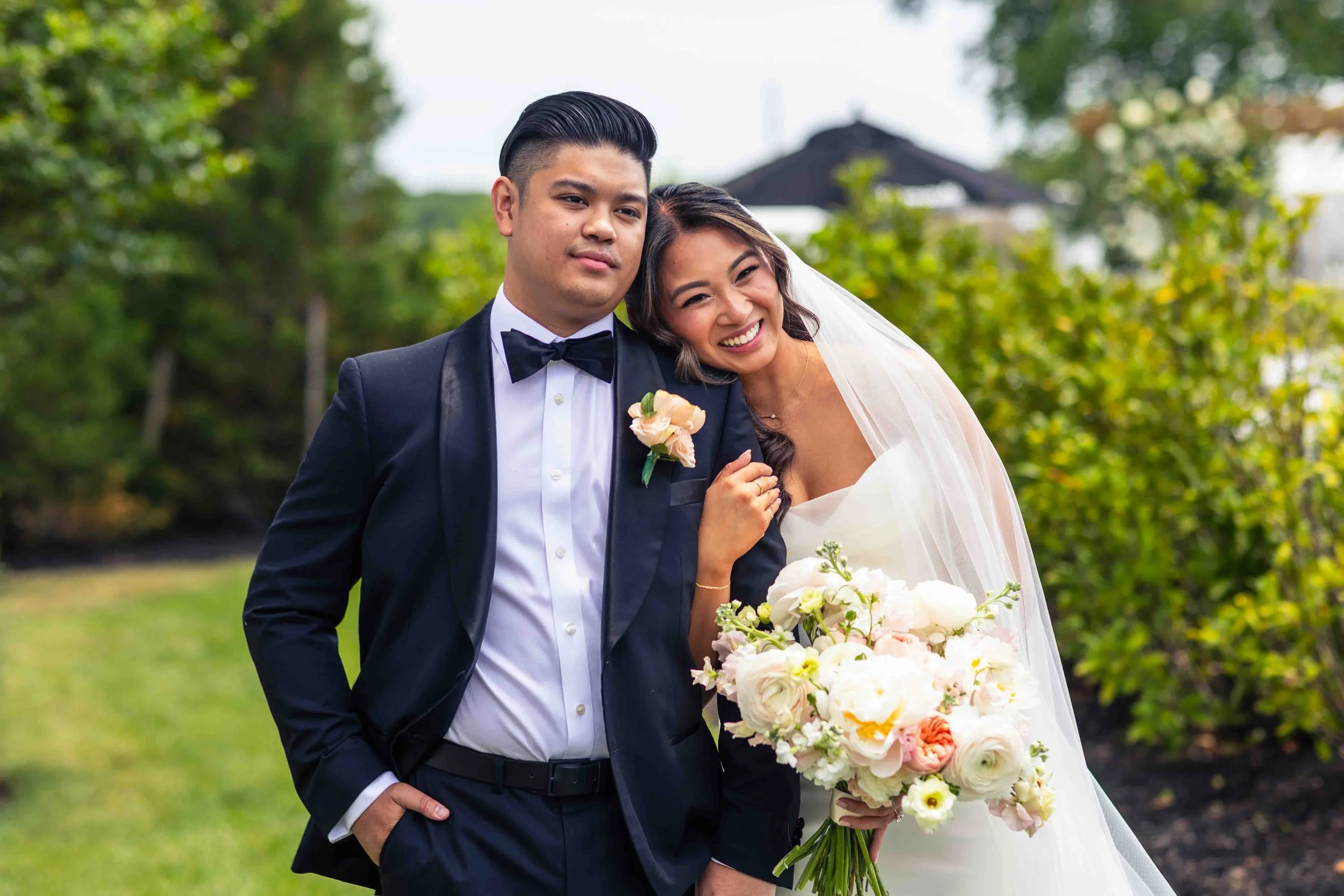 A bride and groom pose outdoors, the bride holding a bouquet and leaning her head on the groom's shoulder, both dressed in wedding attire, with greenery and trees in the background.