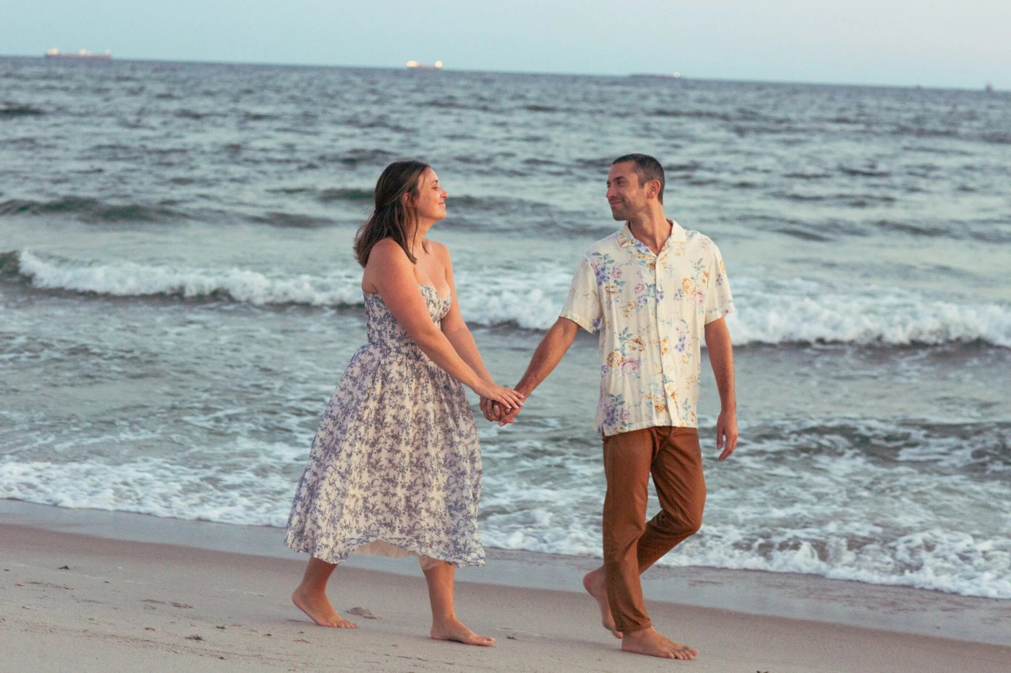 A couple holding hands and walking barefoot along the shoreline of a beach, with ocean waves in the background.
