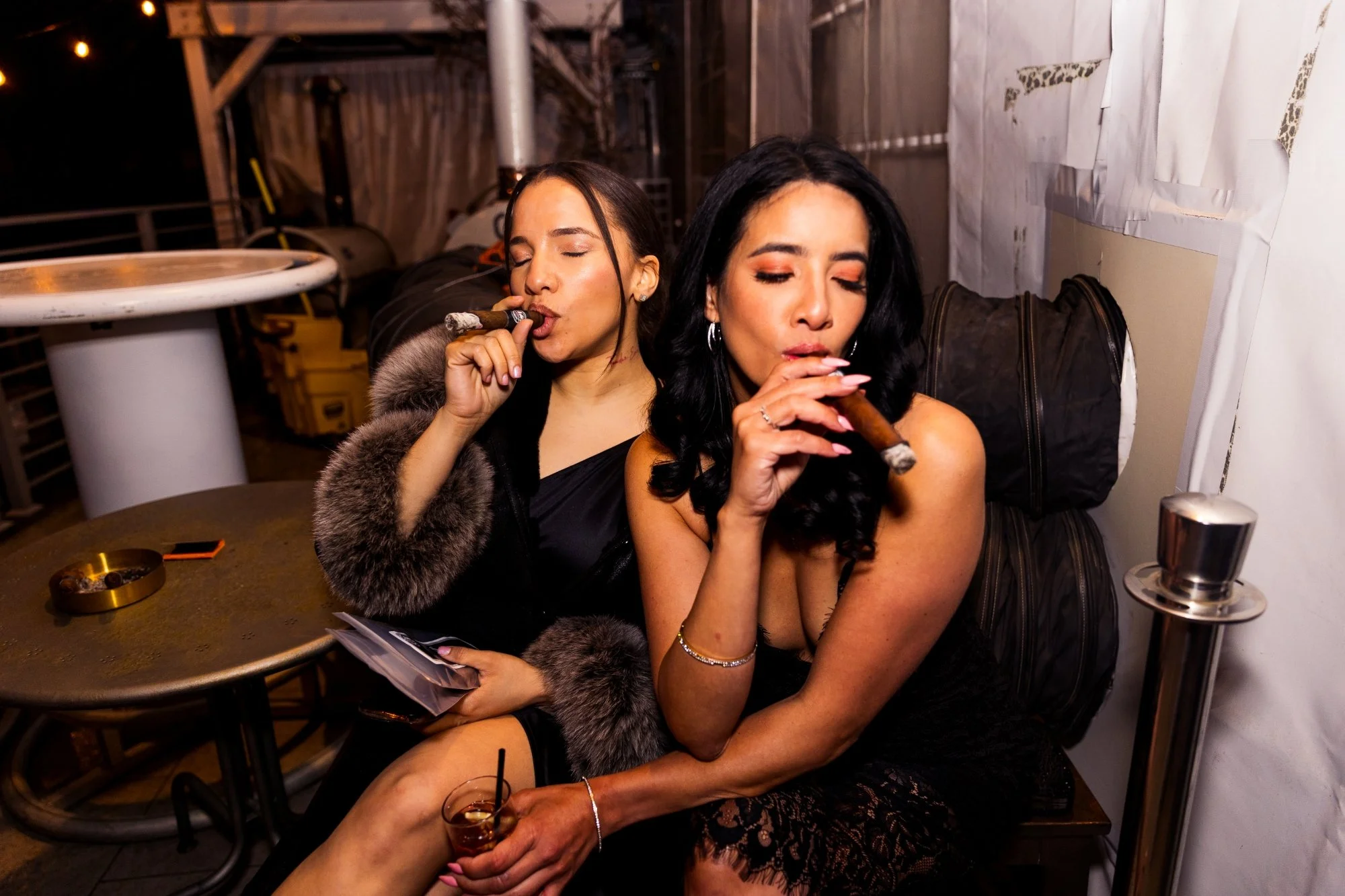 Two women sitting closely together, both smoking cigars, in a dimly lit setting, with the woman in the front holding a cocktail.
