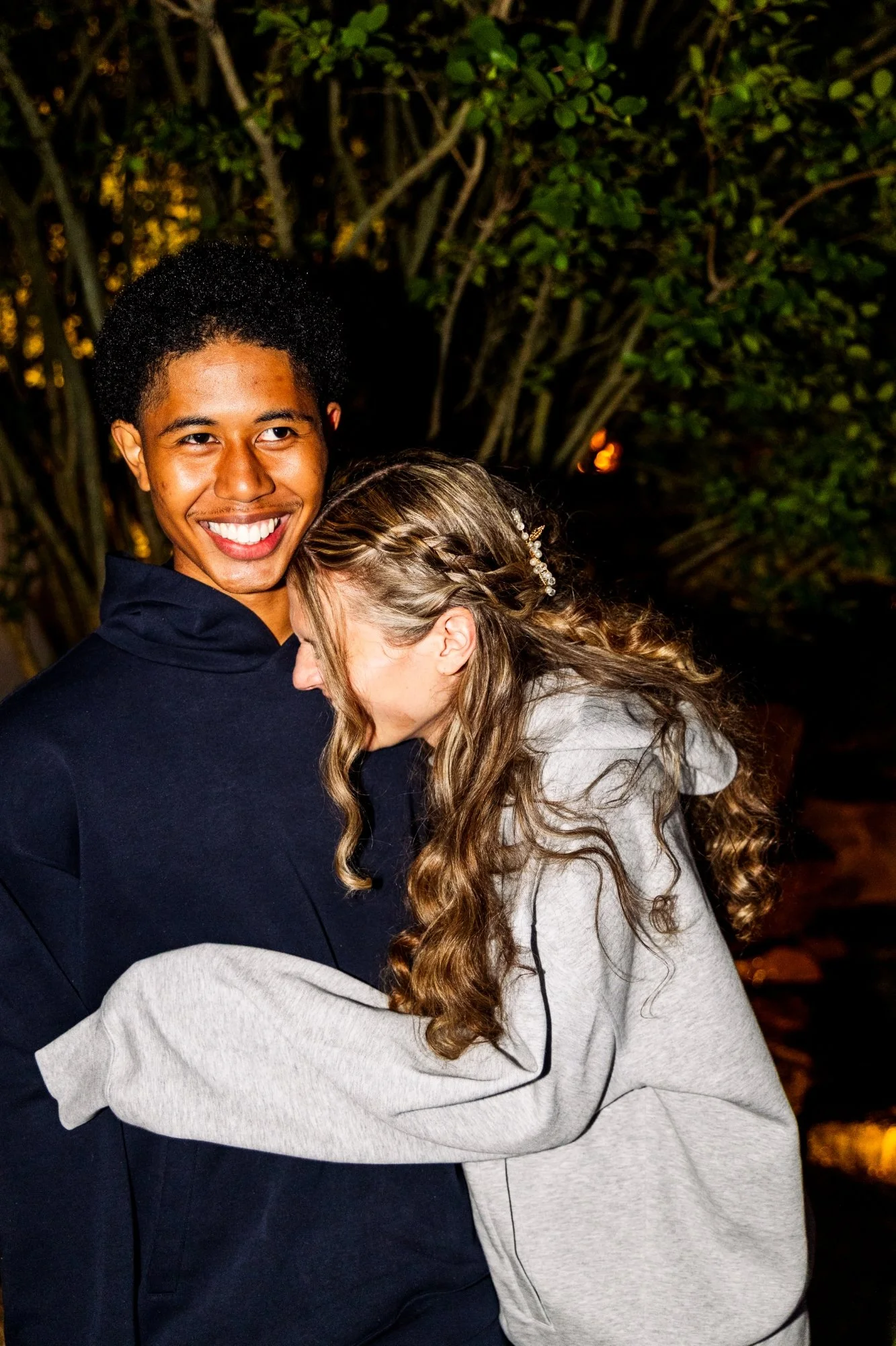 A young man smiling and embracing a woman who is resting her head on his shoulder outdoors at night, with trees and warm lights in the background.