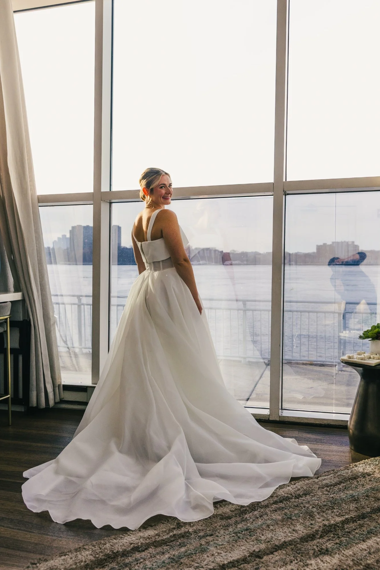 A woman in a wedding dress standing inside near large windows with a view of a river and city skyline, smiling at the camera.