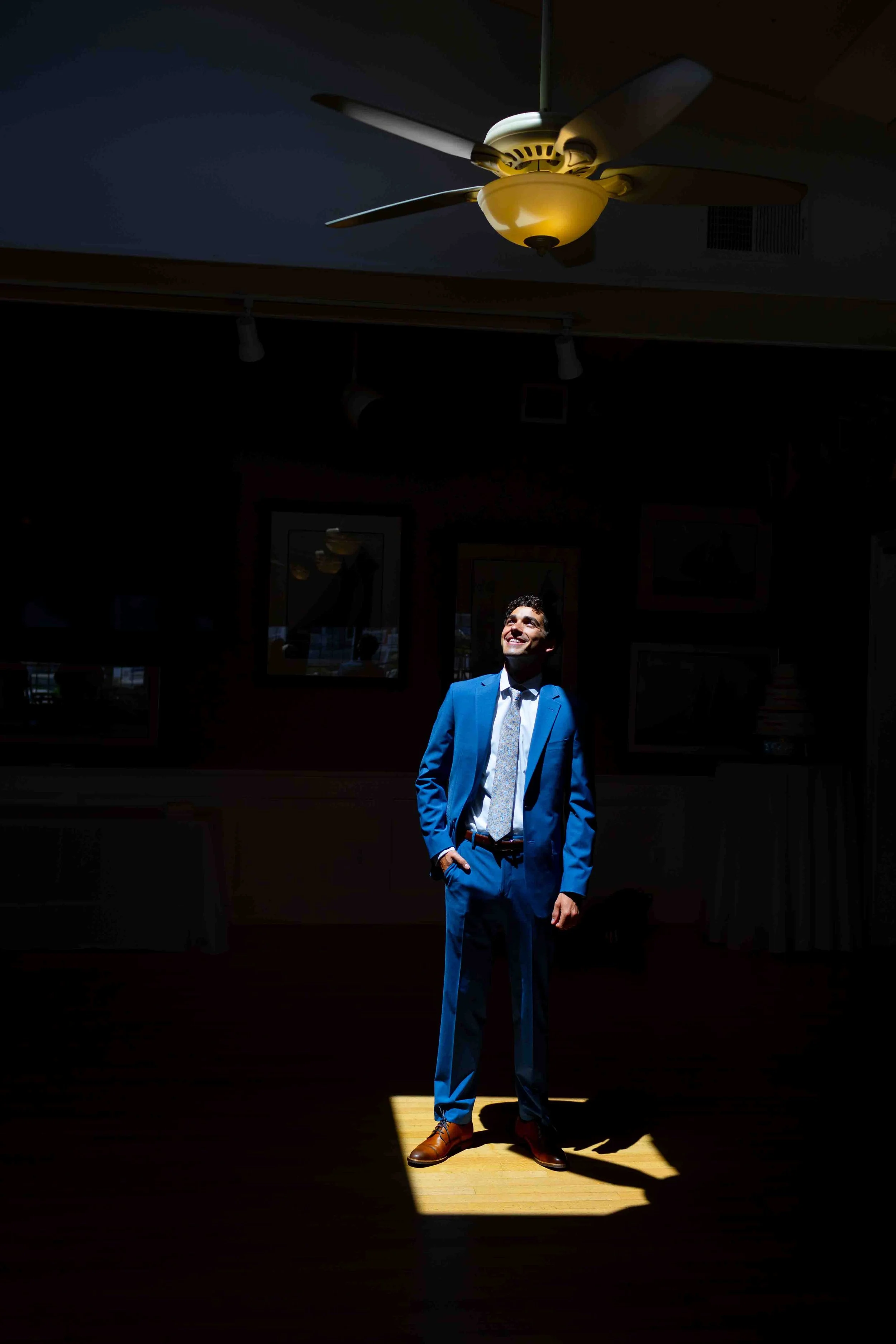 A man in a vibrant blue suit, white shirt, and patterned tie stands in a spotlight with a broad smile, hands in his pockets, in a dimly lit room with artwork on the walls. Overhead ceiling fan is visible.