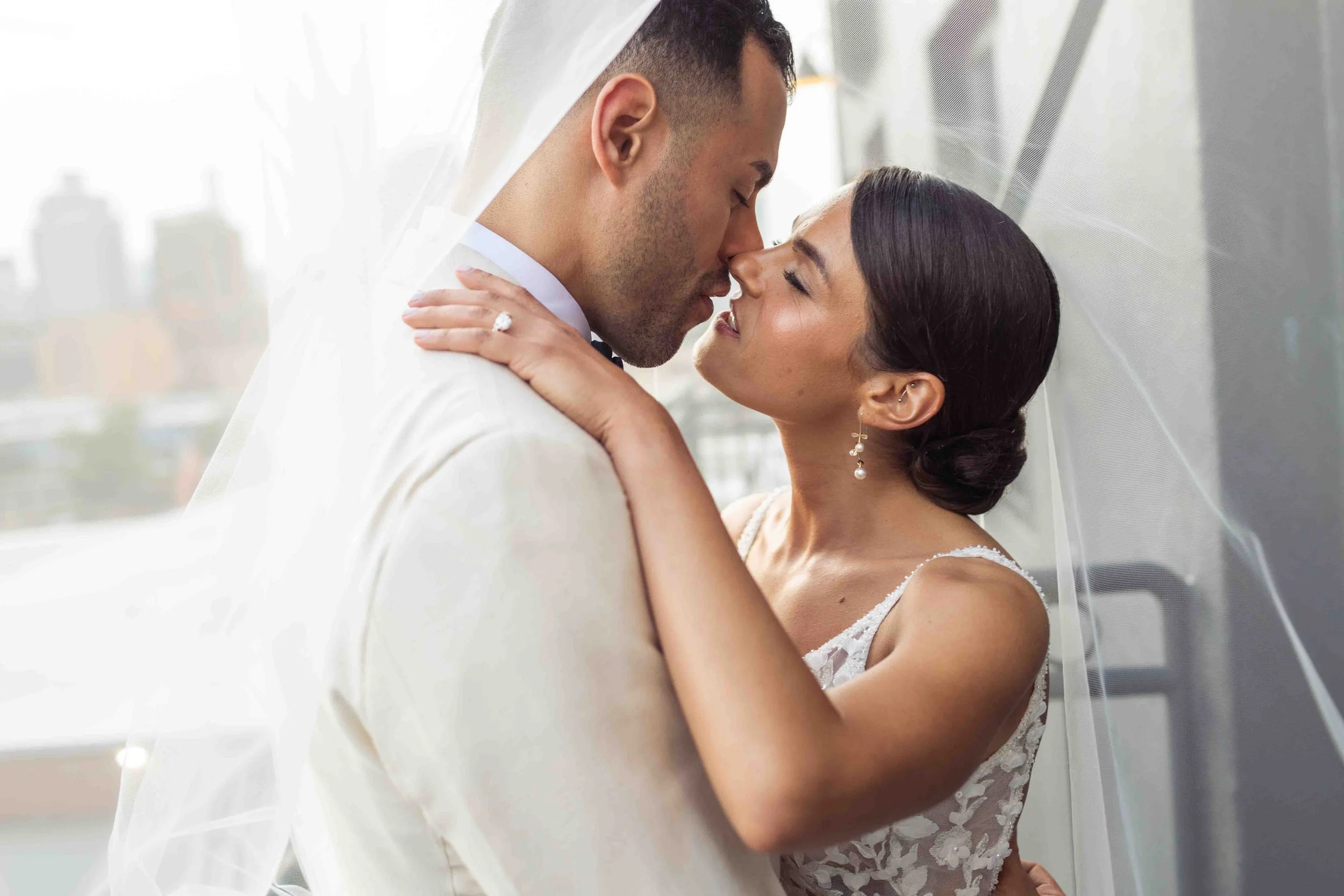 A newlywed couple sharing an intimate moment, with the groom wearing a white suit and veil, and the bride in a white lace dress with earrings, standing in front of a window with city buildings in the background.
