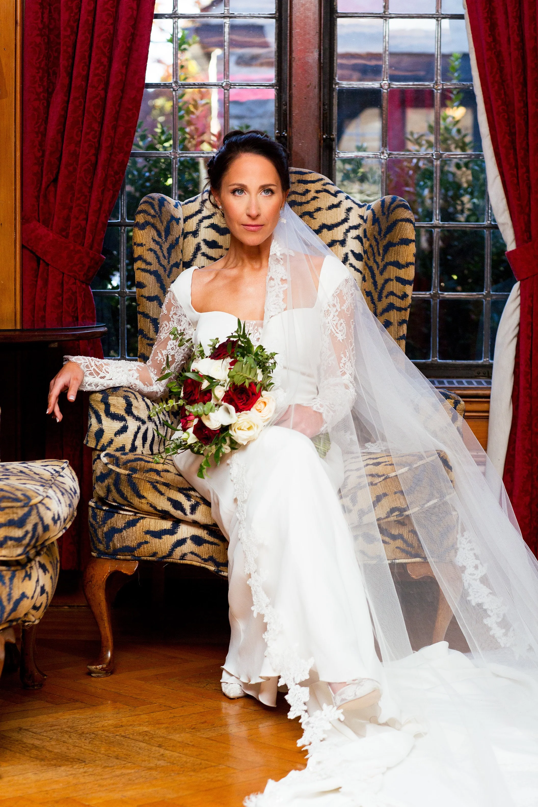 A bride sitting on a tiger-striped armchair inside a room with large windows and red curtains. She is wearing a white wedding dress with lace sleeves and a long veil, holding a bouquet of red, white, and cream flowers.