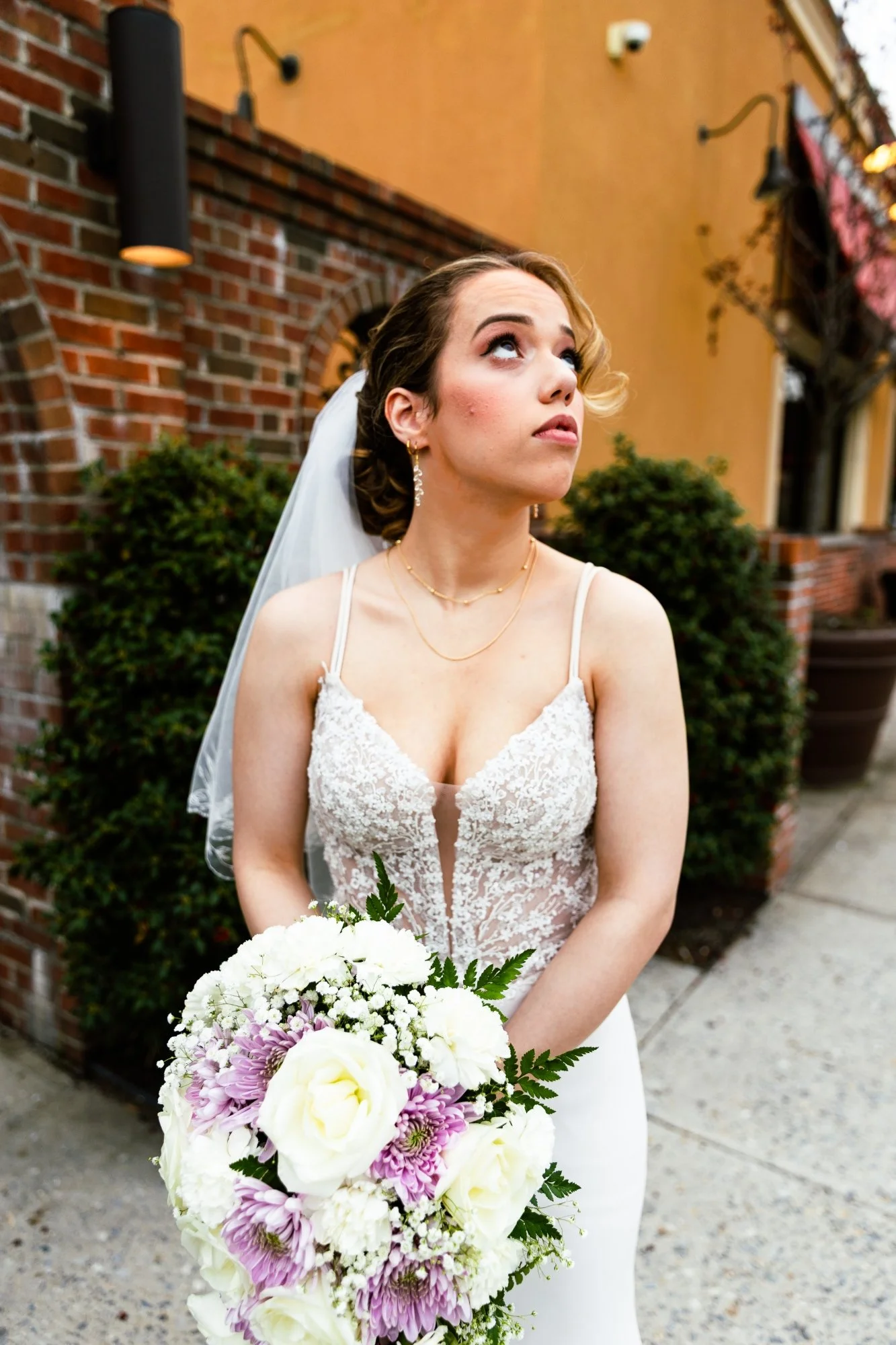 Bride with a wedding dress and veil holding a bouquet of white, pink, and purple flowers standing outdoors near a brick building.