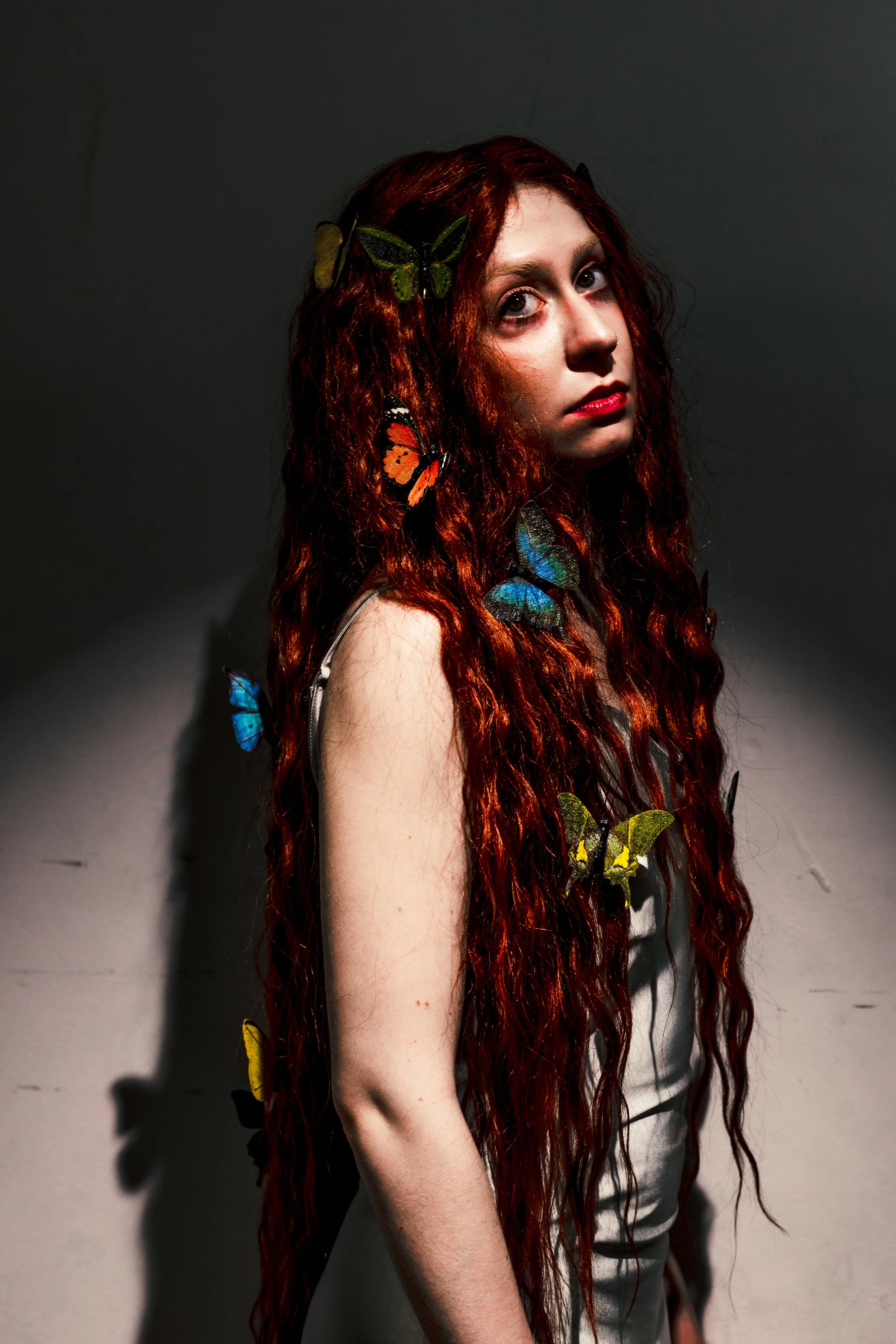 Person wearing a black robe with red floral embroidery, standing against a textured dark background, hands near head, looking to the side.