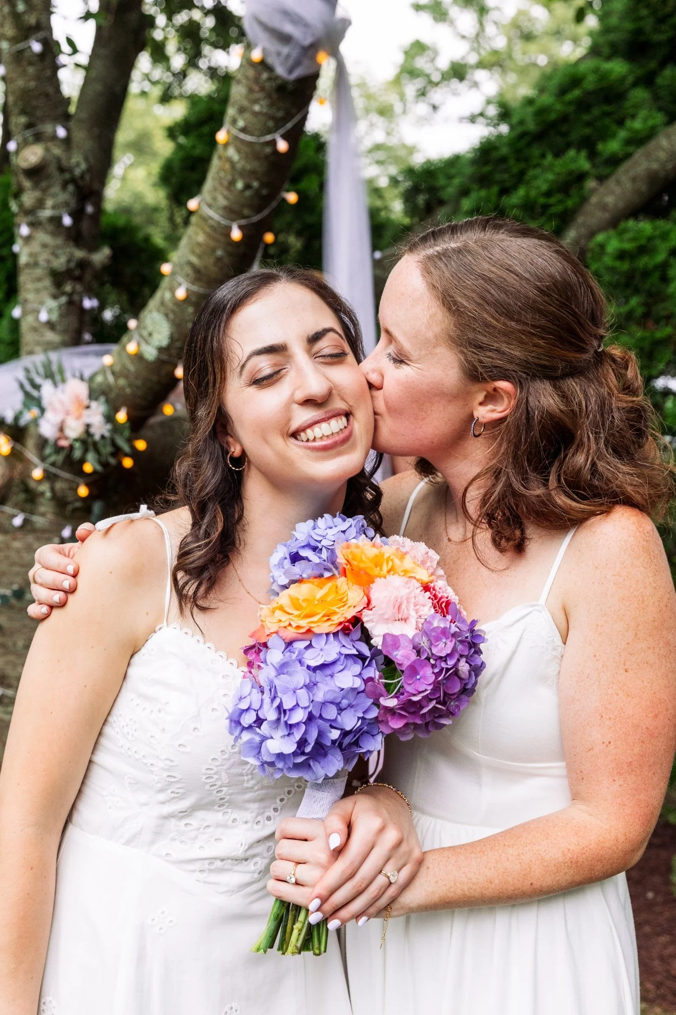 Two women, one kissing the other on the cheek, holding a bouquet of colorful hydrangeas and roses, with a decorated outdoor tree backdrop in a garden setting.