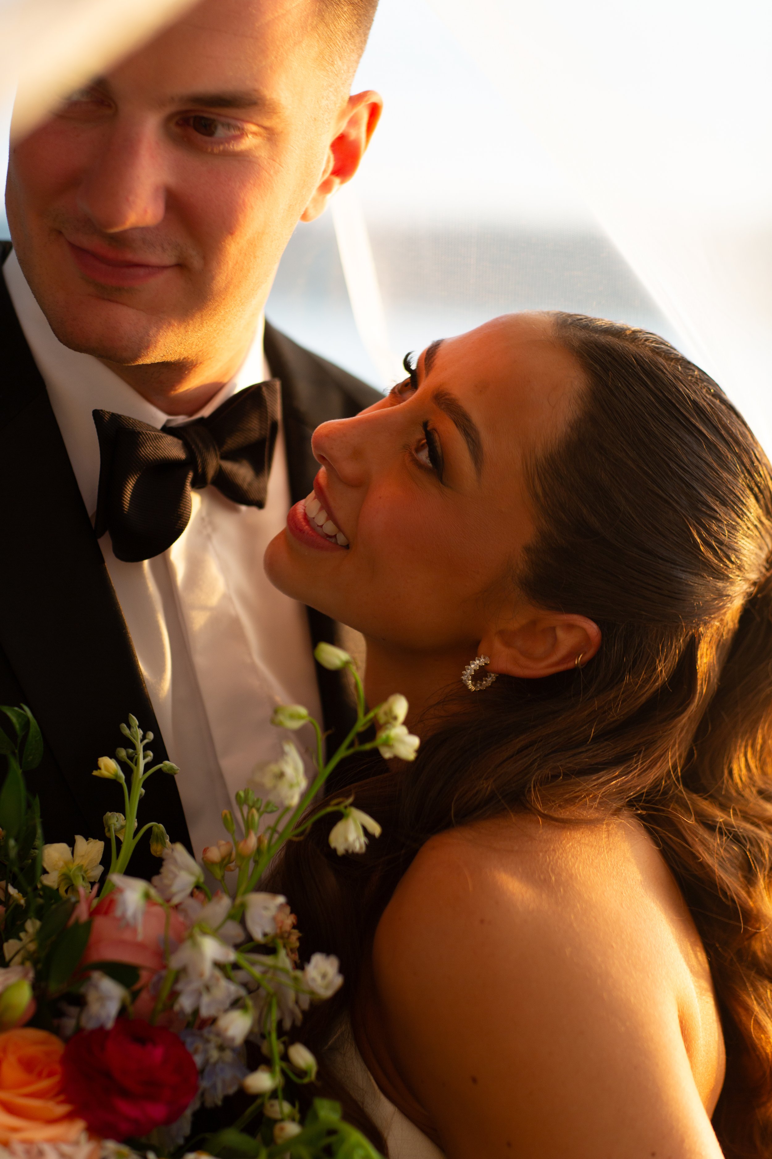 A smiling woman with long brown hair, earrings, and a white dress hugging a man in a tuxedo, holding a bouquet of flowers during a wedding.