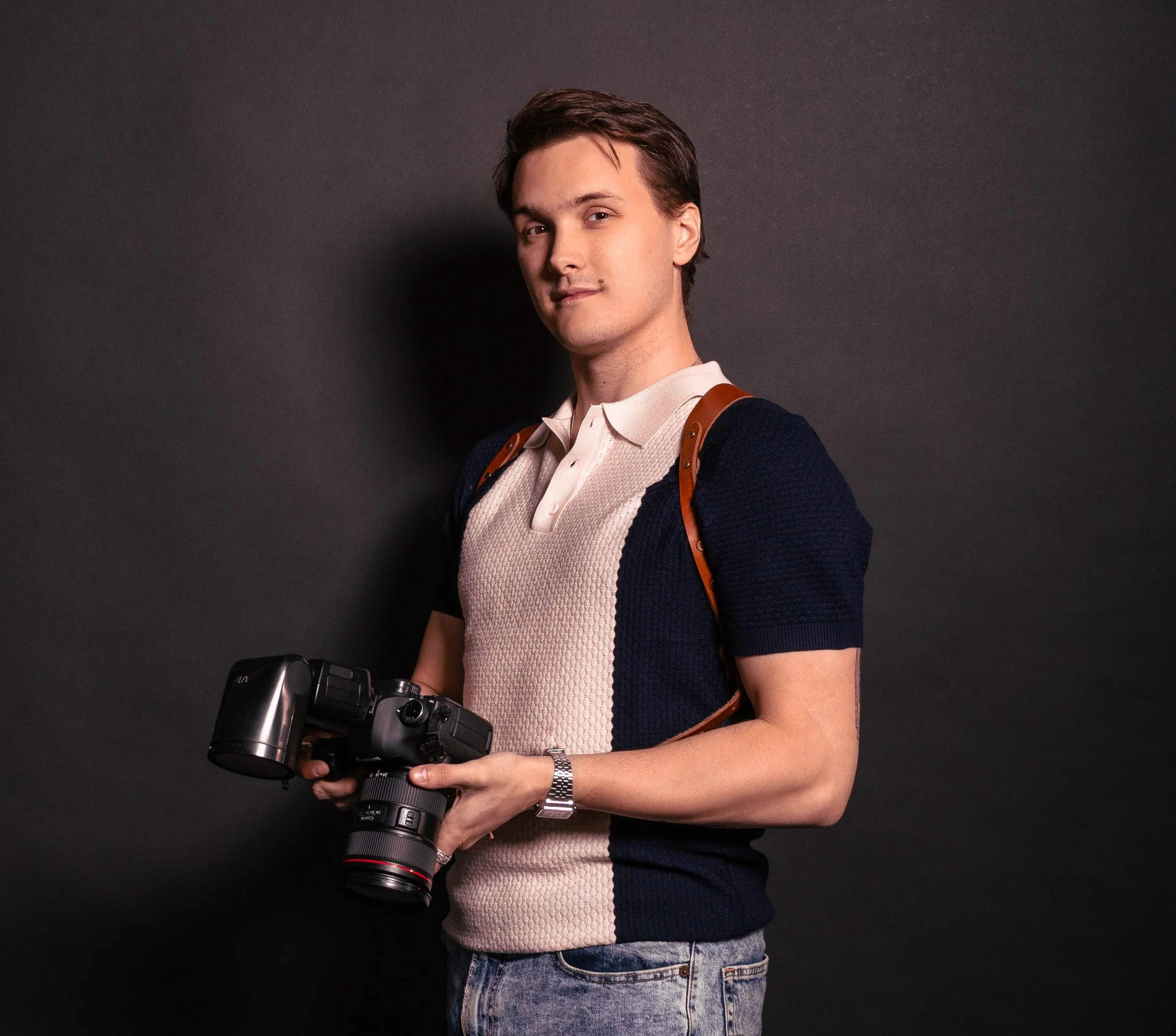 Young man with brown hair holding a professional camera, wearing a beige and navy blue short-sleeved shirt, jeans, and a watch, standing against a dark background.
