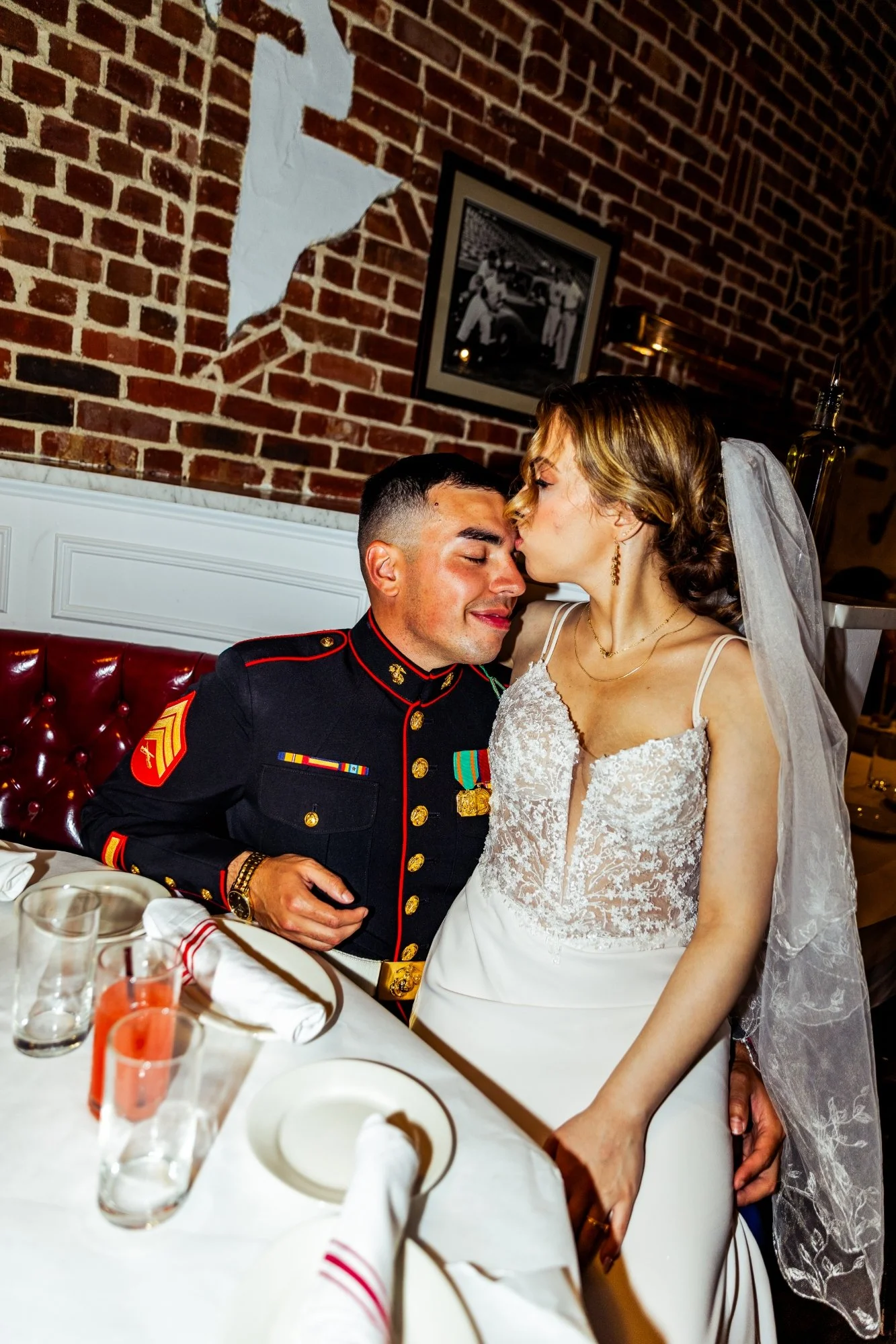 A bride and groom sharing a kiss at a wedding reception. The groom is dressed in a military uniform, and the bride is wearing a lace wedding gown and veil.
