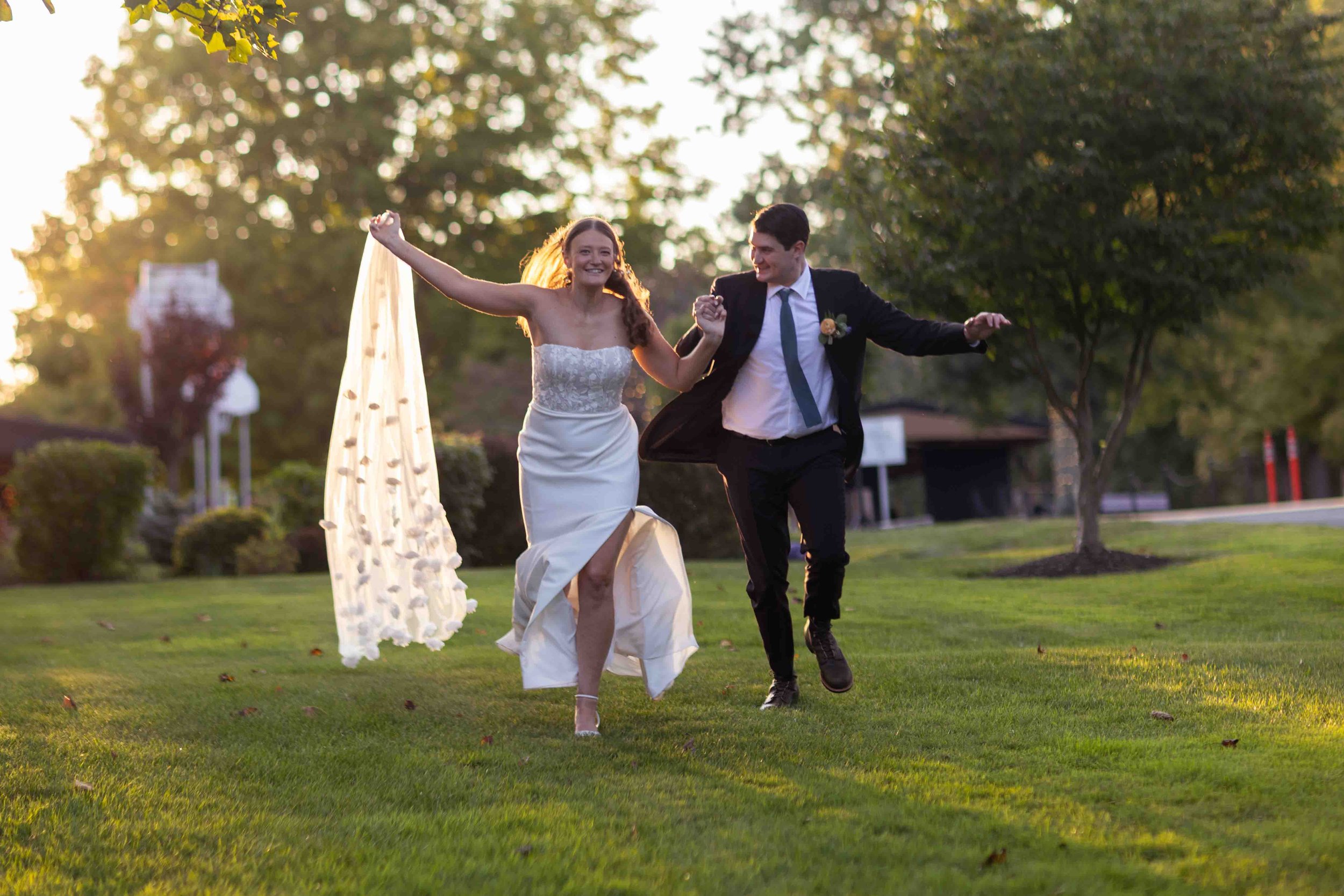 A newlywed couple in wedding attire joyfully running in a grassy park during sunset, smiling and holding hands.