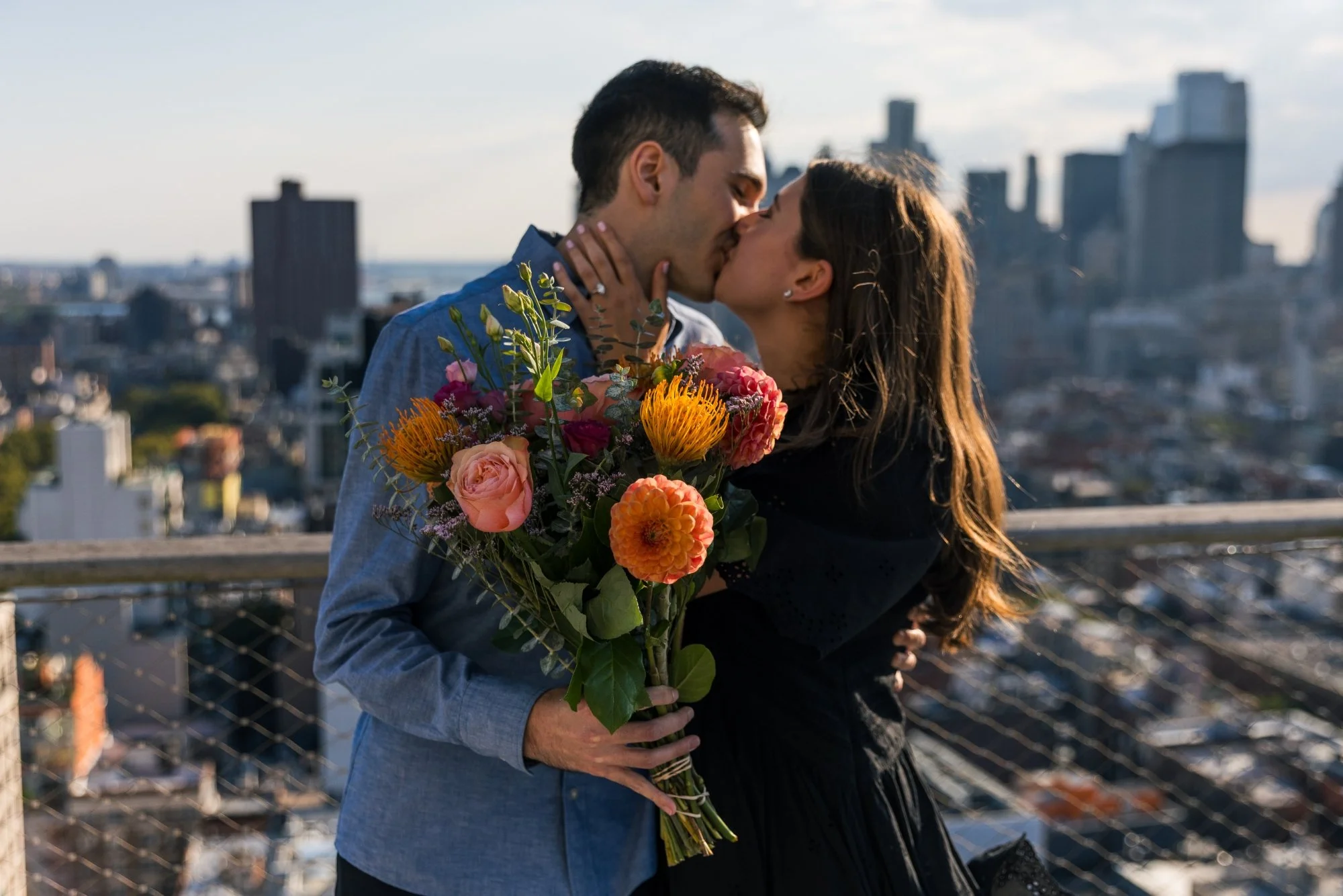 A couple sharing a kiss on a city rooftop with a bouquet of flowers, city buildings in the background.