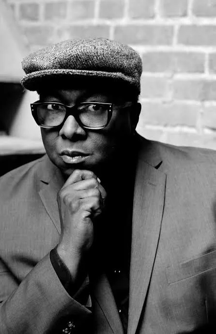 Black-and-white portrait of Music Director, Abdul Hamid Royal, a flat cap, and a suit jacket, resting his chin on his hand.