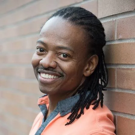 Choreographer, Byron Easley, wearing a coral shirt, posing against a brick wall