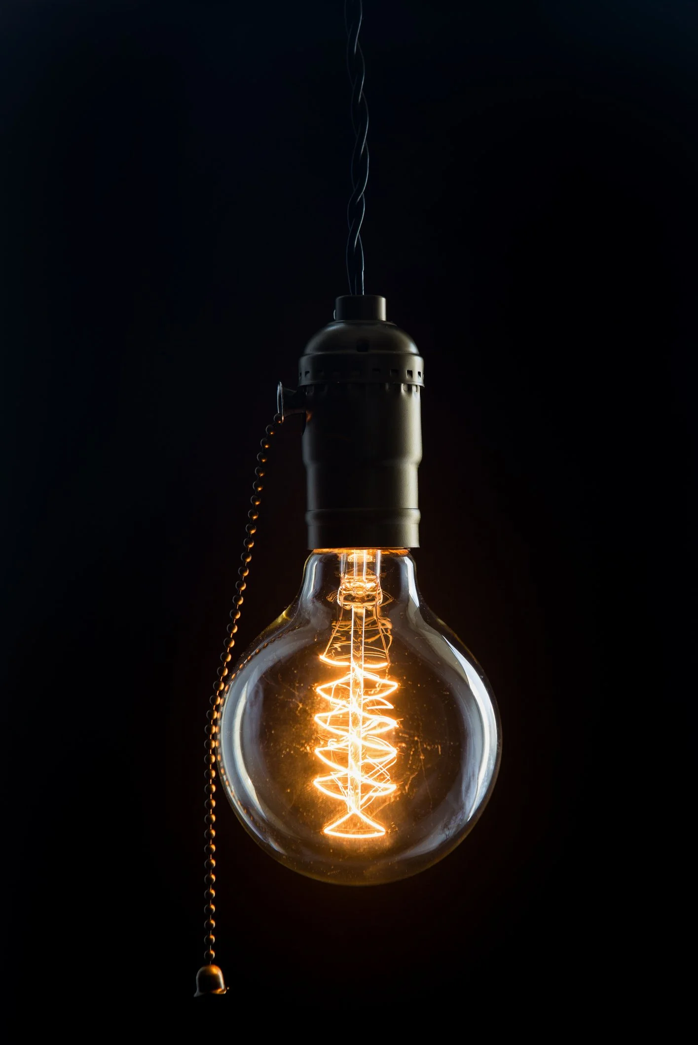 Glowing vintage Edison light bulb hanging against a black background with a pull chain.