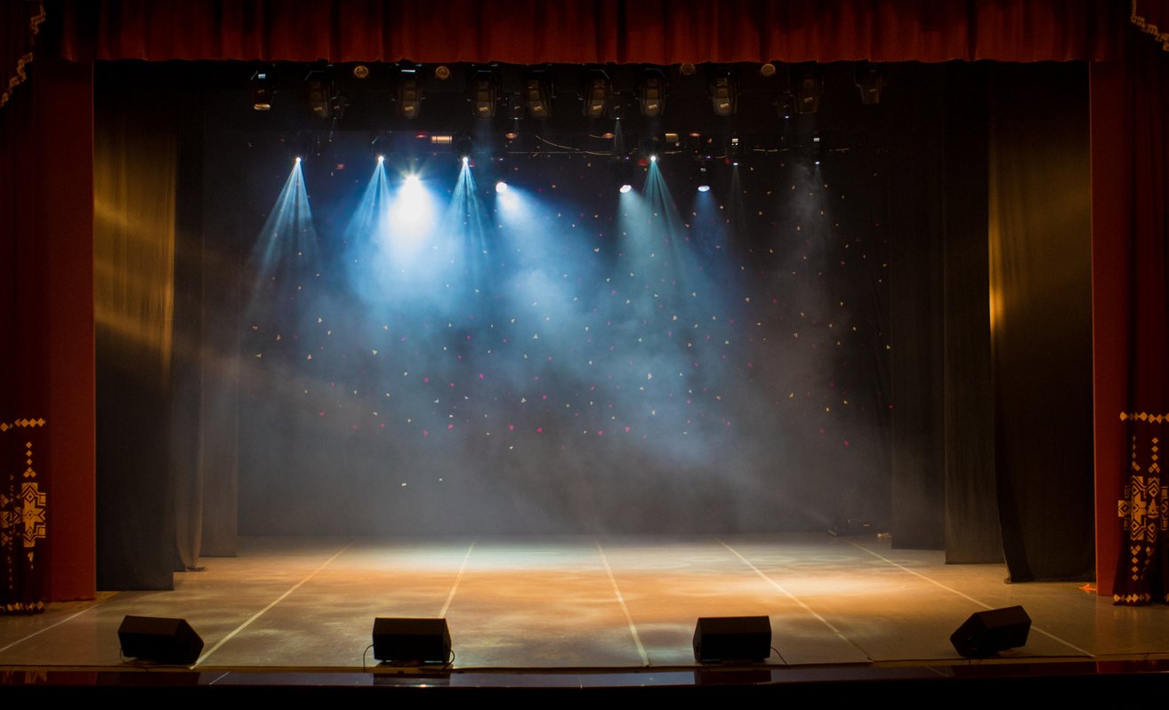 Empty theater stage with red curtains, blue spotlights, haze, and black floor speakers