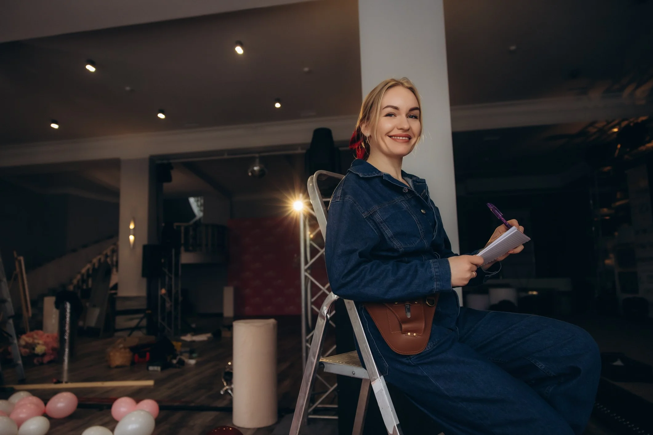 Smiling stage manager in a denim jumpsuit writing in a notebook while seated on a step ladder backstage