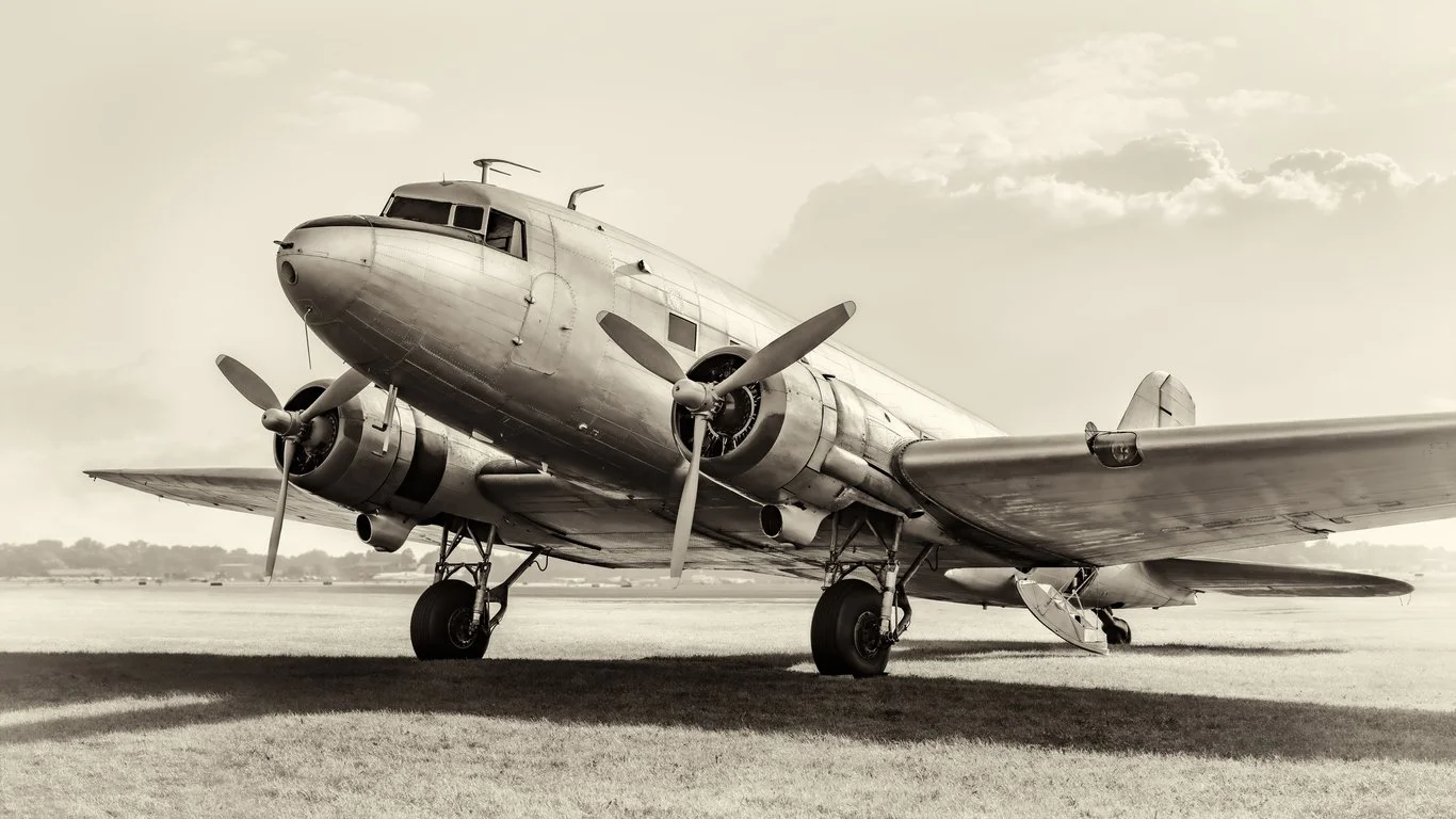 Sepia-toned vintage twin-engine propeller airplane parked on an airfield beside the water.