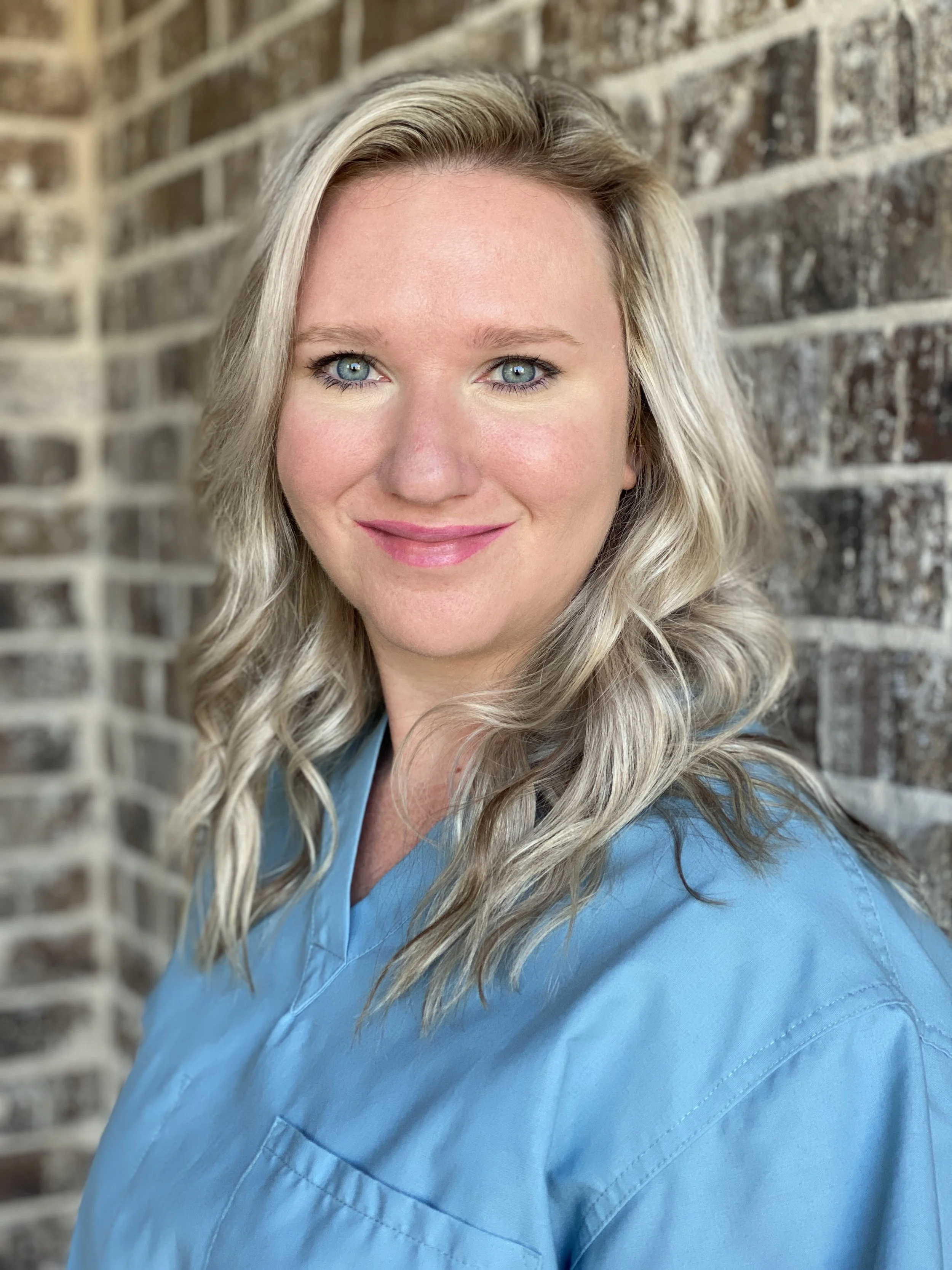A smiling female nurse practitioner with blonde hair and blue eyes smiling in front of a brick wall, wearing blue scrubs.