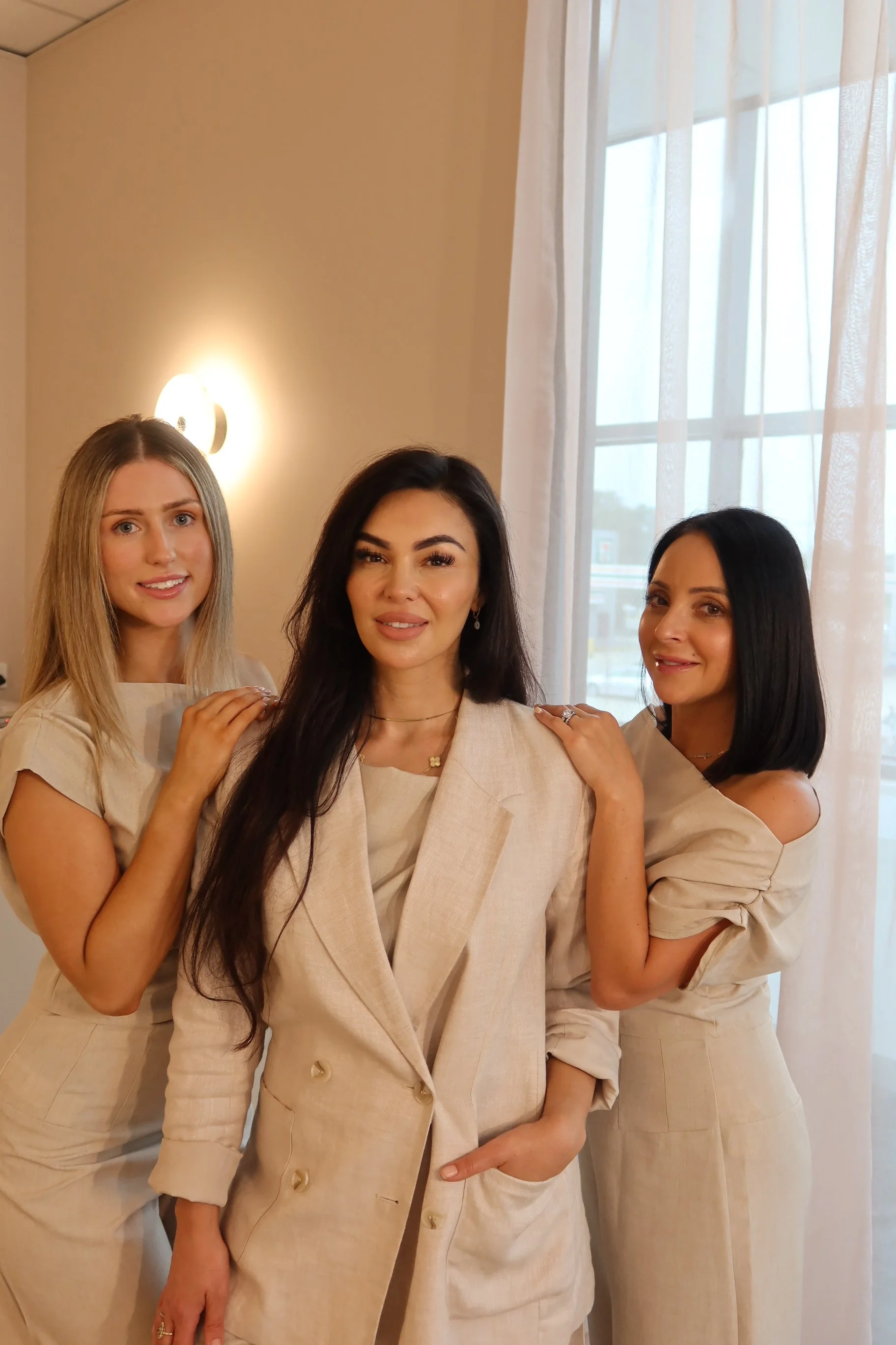 Three women in beige outfits posing together indoors, beige wall with a wall sconce, and a window with white curtains in the background.