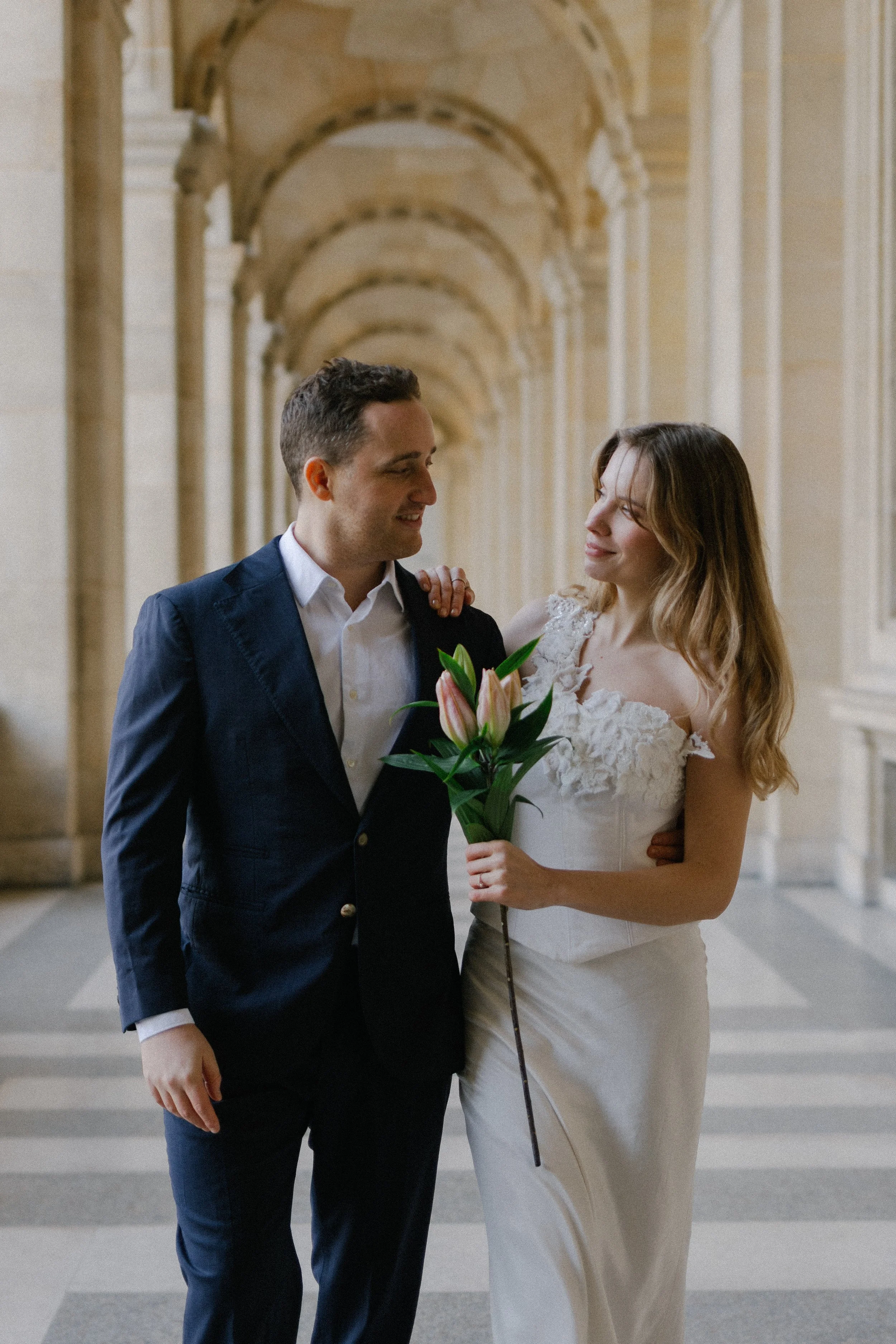 Couple eloping in the Louvre museum together smiling to eachother