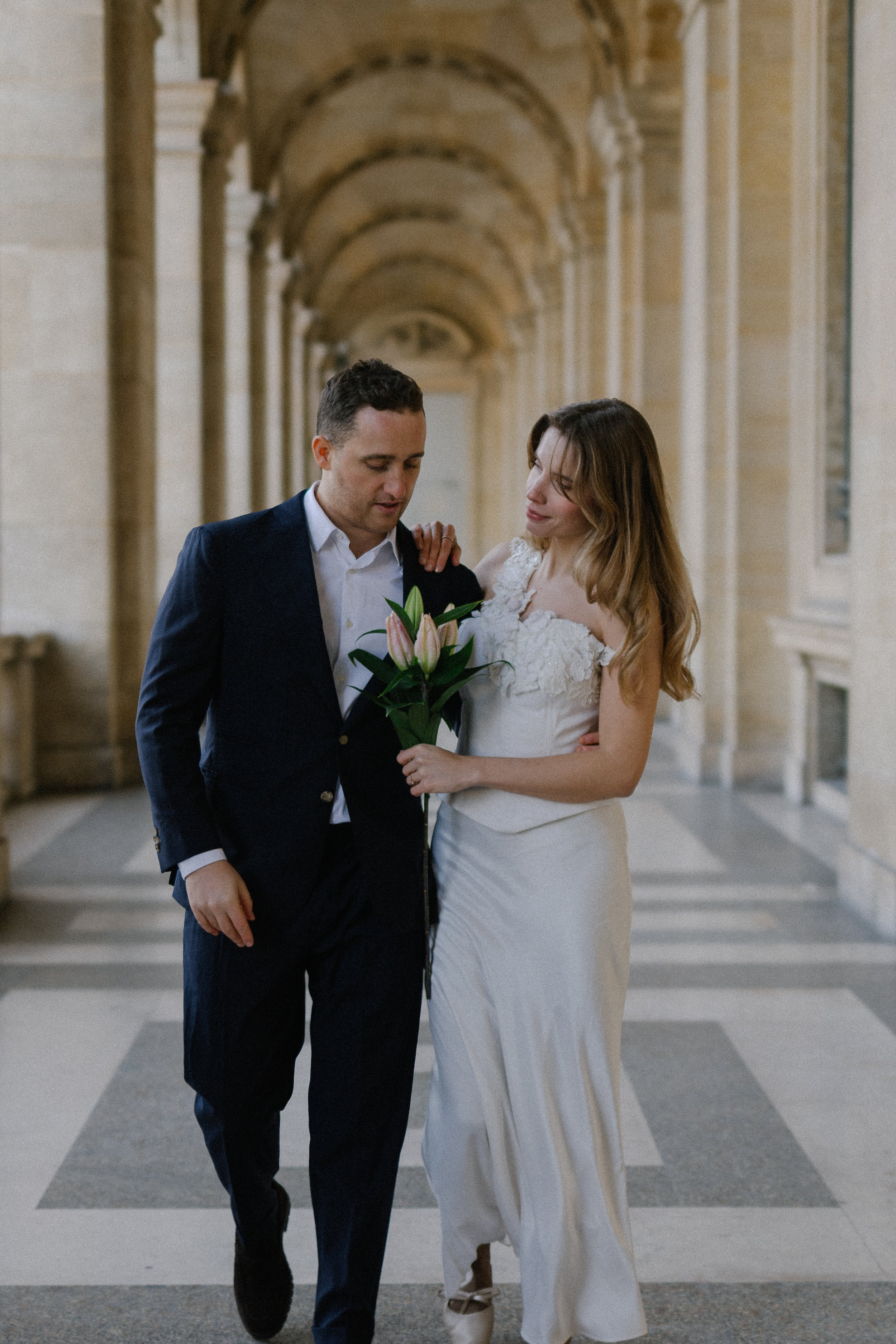 Bride and groom walking together in the louvre museum, with flowers in their hands, an emotive look in their face, being happy to take photography in their best day of their lives.