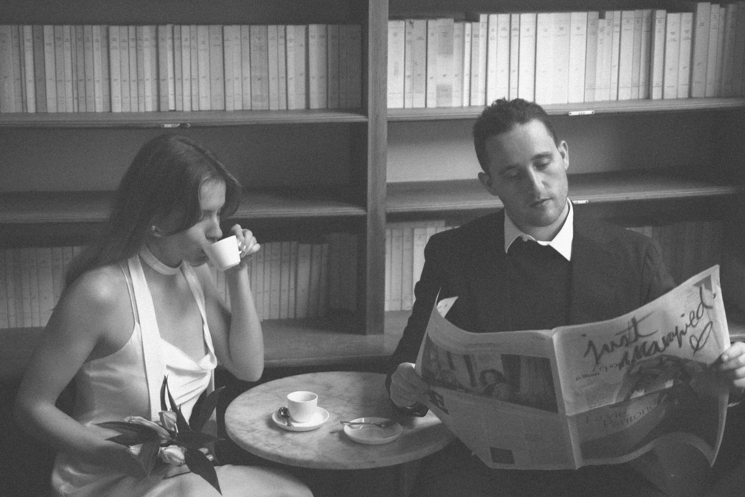 A black-and-white photo of a woman drinking coffee at a small table and a man reading a newspaper in a room with bookshelves.