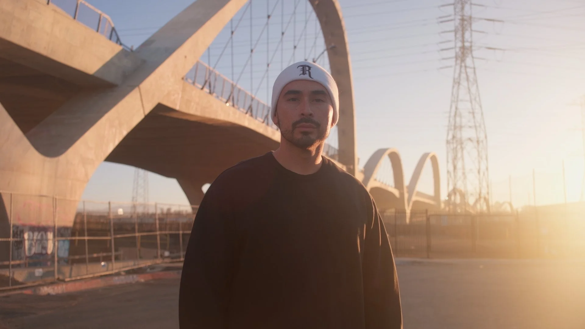 A man standing outdoors near an arched bridge during sunset, wearing a white beanie and a black shirt.