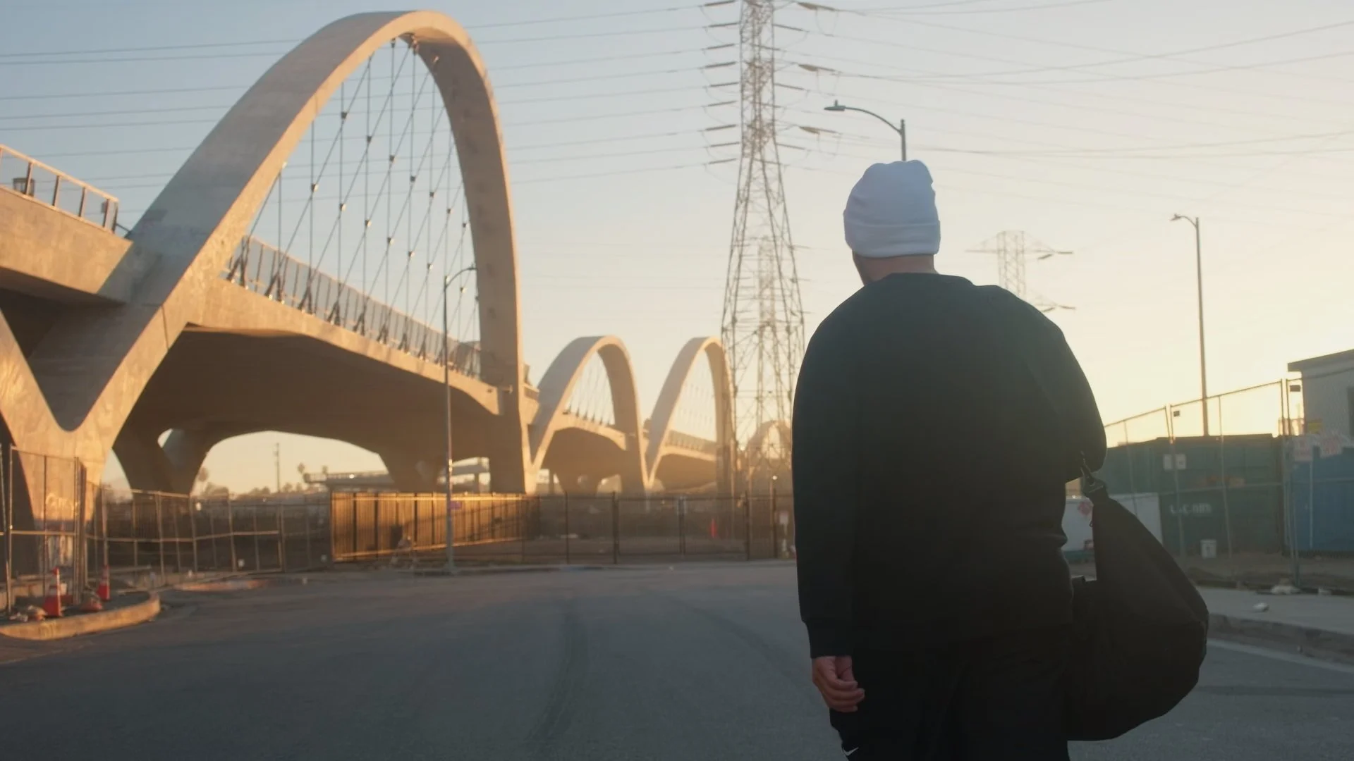 A person wearing a white beanie and black jacket with a backpack standing near a bridge and electrical towers during sunset or sunrise.