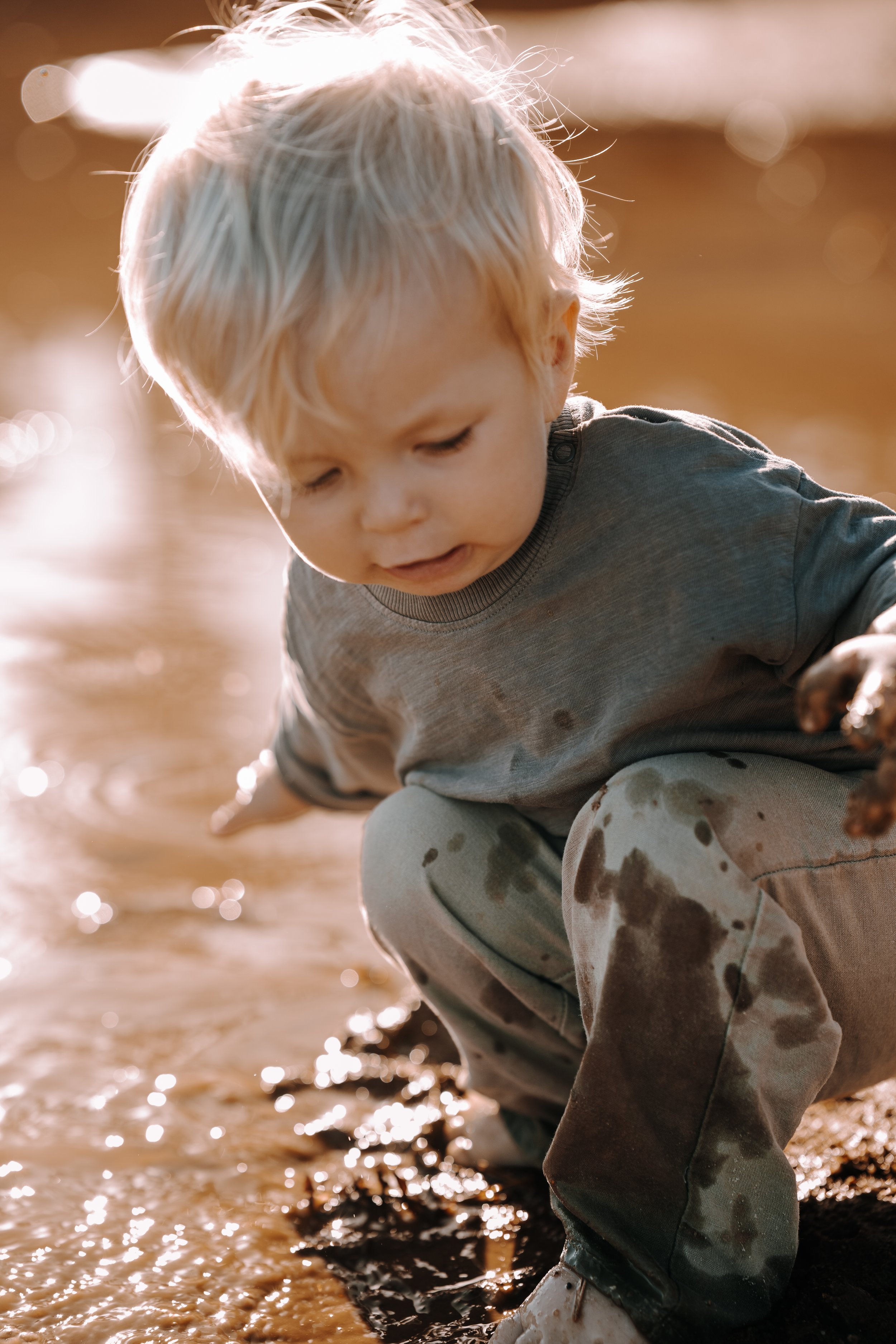 Junge mit blondem Haar, der im Wasser sitzt und nach unten schaut, Sonnenlicht reflektiert auf Wasser und Sand, trug eine graue Kleidung mit braunen Flecken.