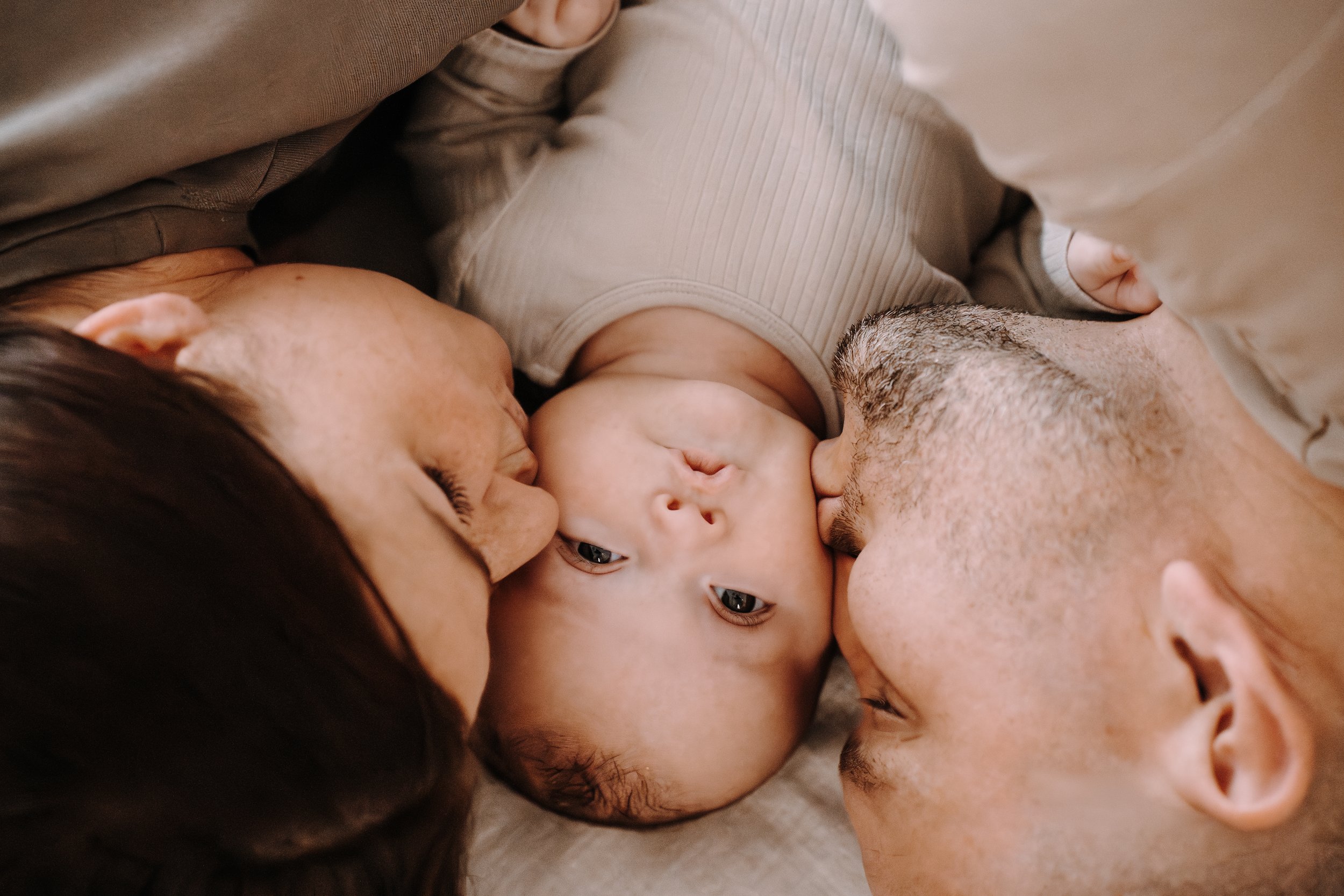 Drei Menschen, zwei Erwachsene und ein Baby, liegen auf dem Boden und berühren sich die Stirn, während sie sich liebevoll anschauen.
