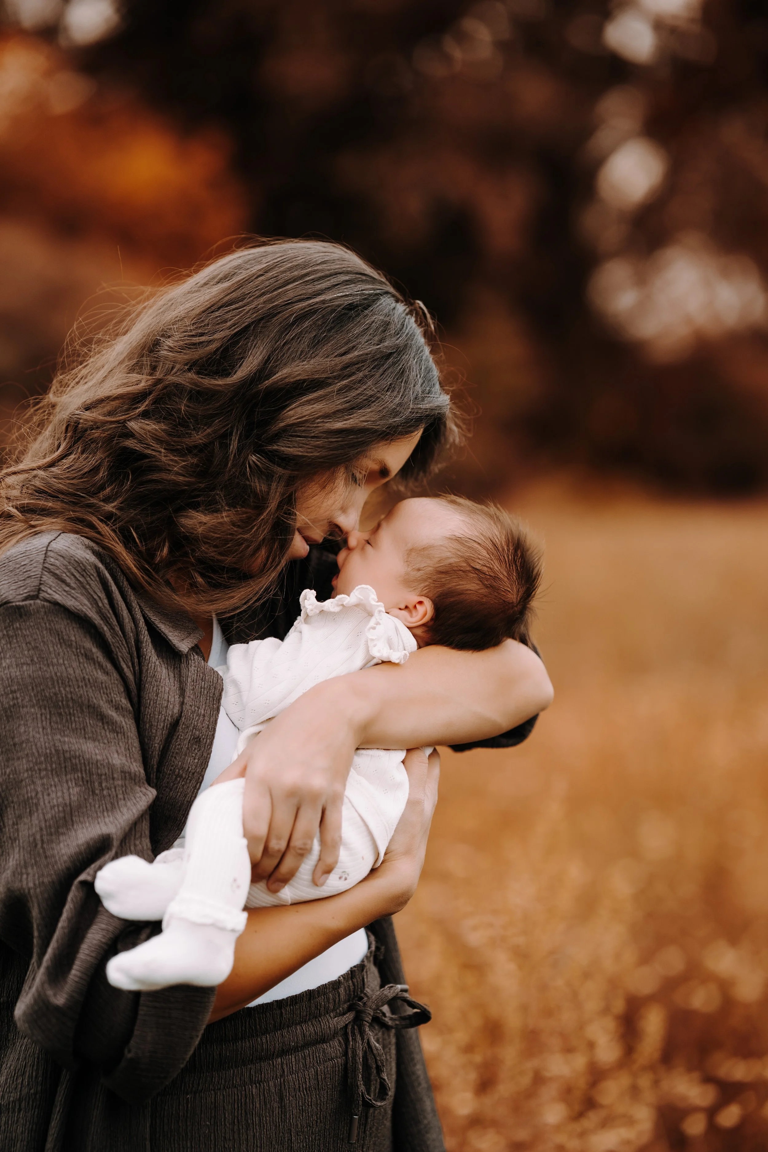 Eine Frau mit braunen, gewellten Haaren hält ein Baby im Arm. Sie sind draußen in einem Herbstwald mit bunten Blättern im Hintergrund.