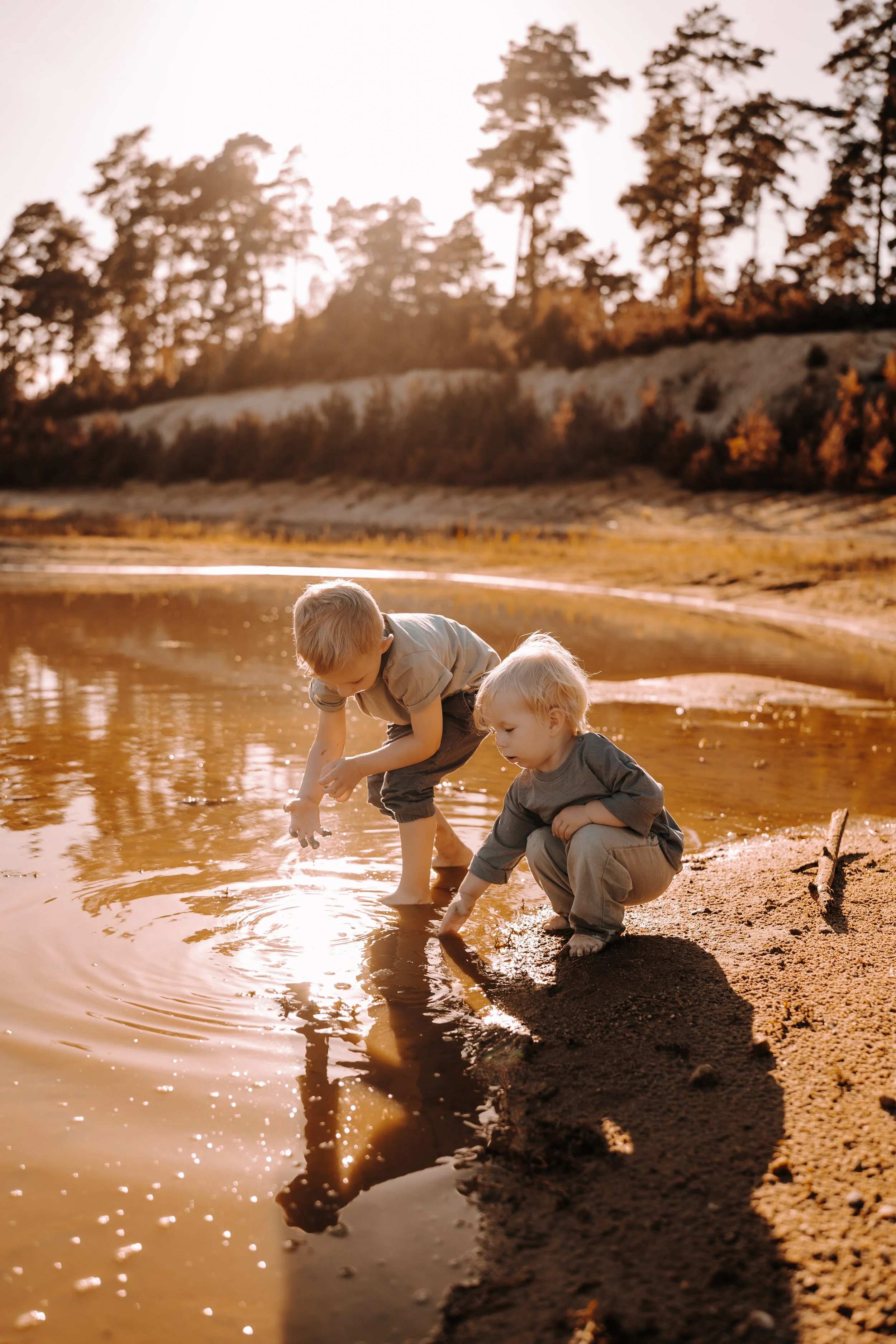 Zwei Kinder spielen am Flussufer bei Sonnenuntergang, sie bücken sich ins Wasser und berühren es mit den Händen, im Hintergrund Bäume und Natur.