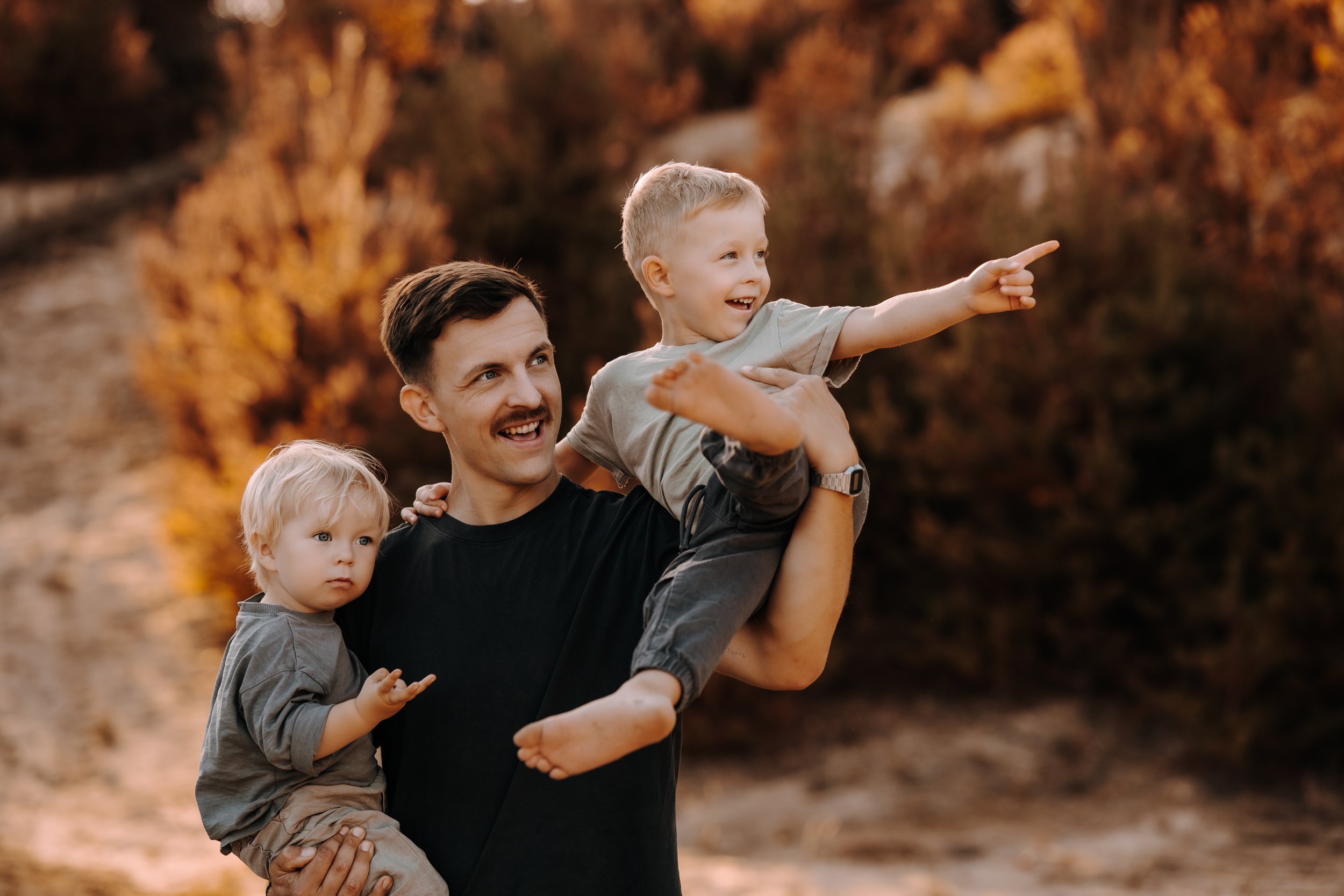 Ein Mann trägt ein lachendes Kind auf den Schultern, während ein weiteres Kind neben ihnen steht und in die Kamera schaut. Es ist Herbst mit bunten Baumblättern im Hintergrund.