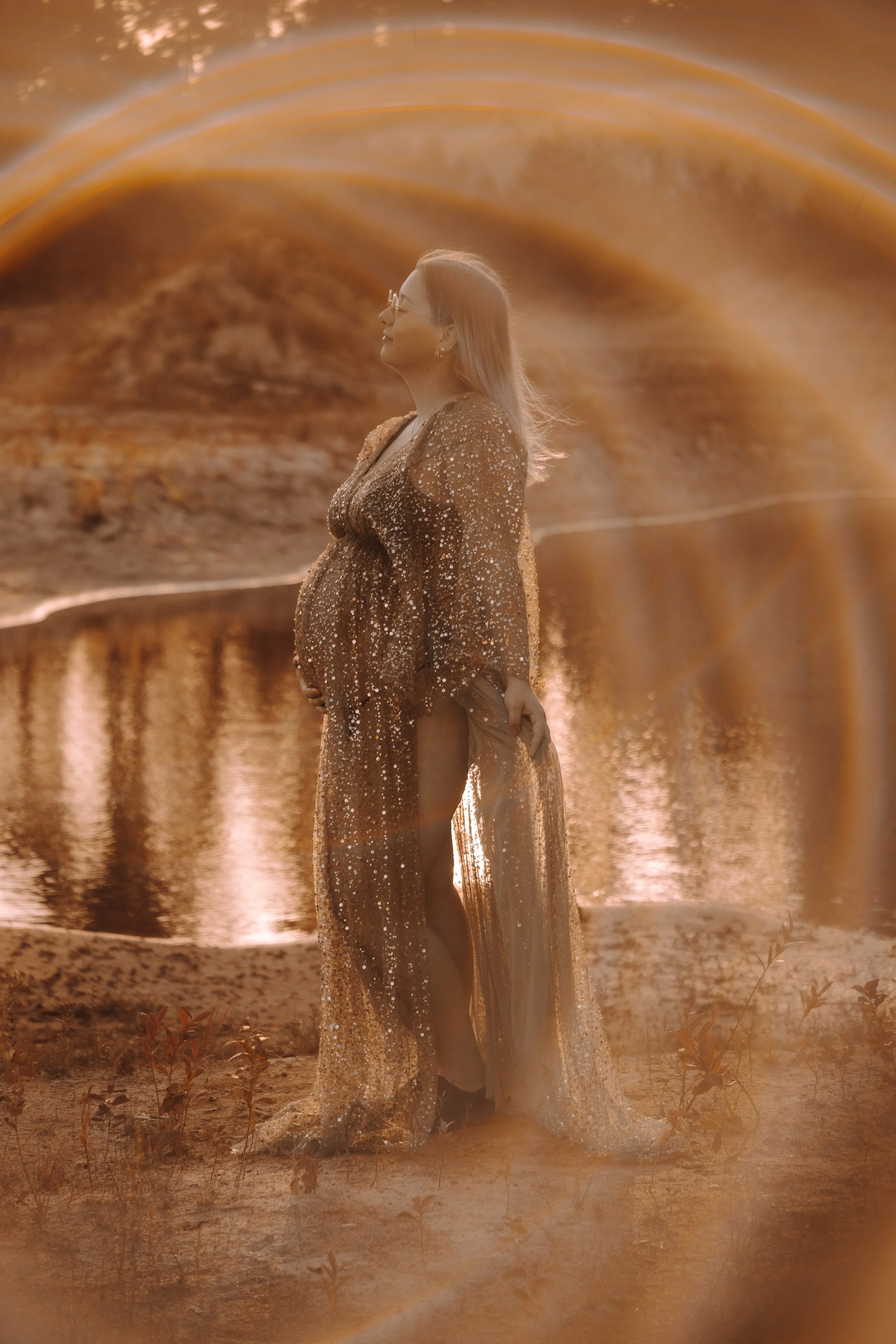 Eine schwangere Frau trägt ein glitzerndes, goldfarbenes Kleid, steht am Ufer eines Sees mit einer goldenen, regenbogenähnlichen Lichtsäule im Himmel im Hintergrund. Sie trägt Brille und schaut nach oben.