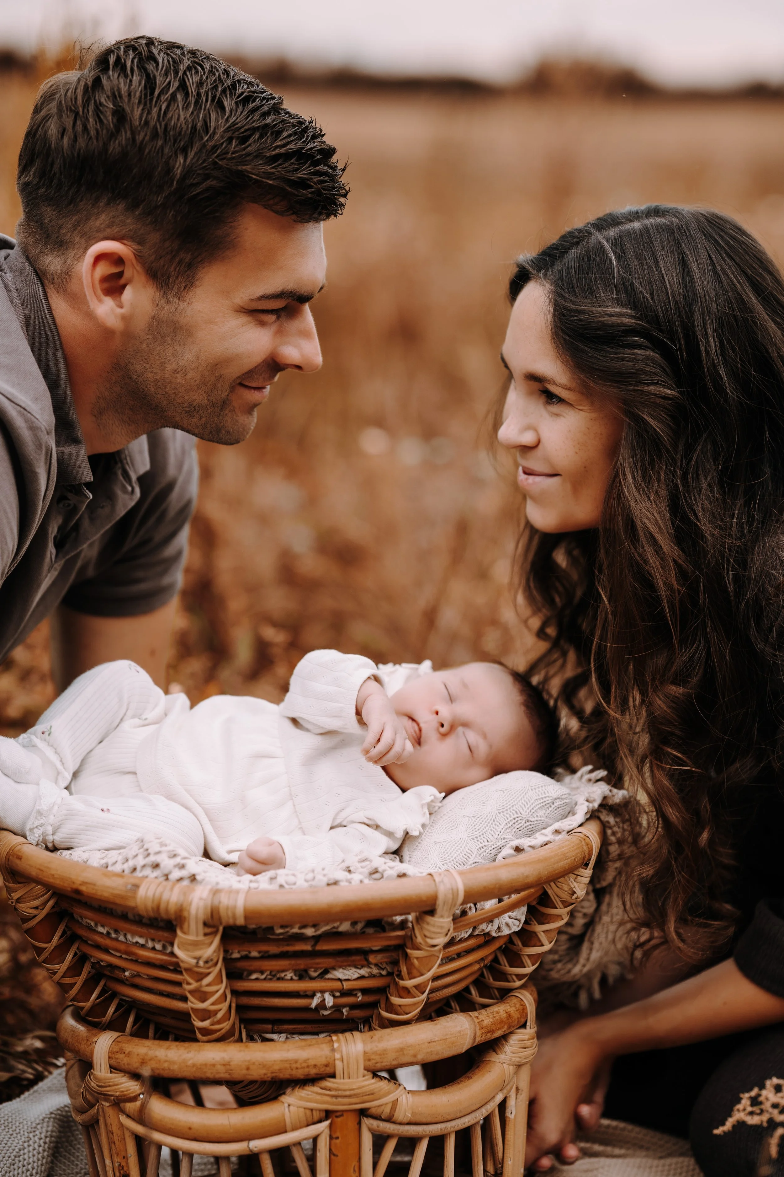 Ein Mann und eine Frau betrachten ein schlafendes Baby in einem Korb, im Freien mit braunem, herbstlichem Hintergrund.